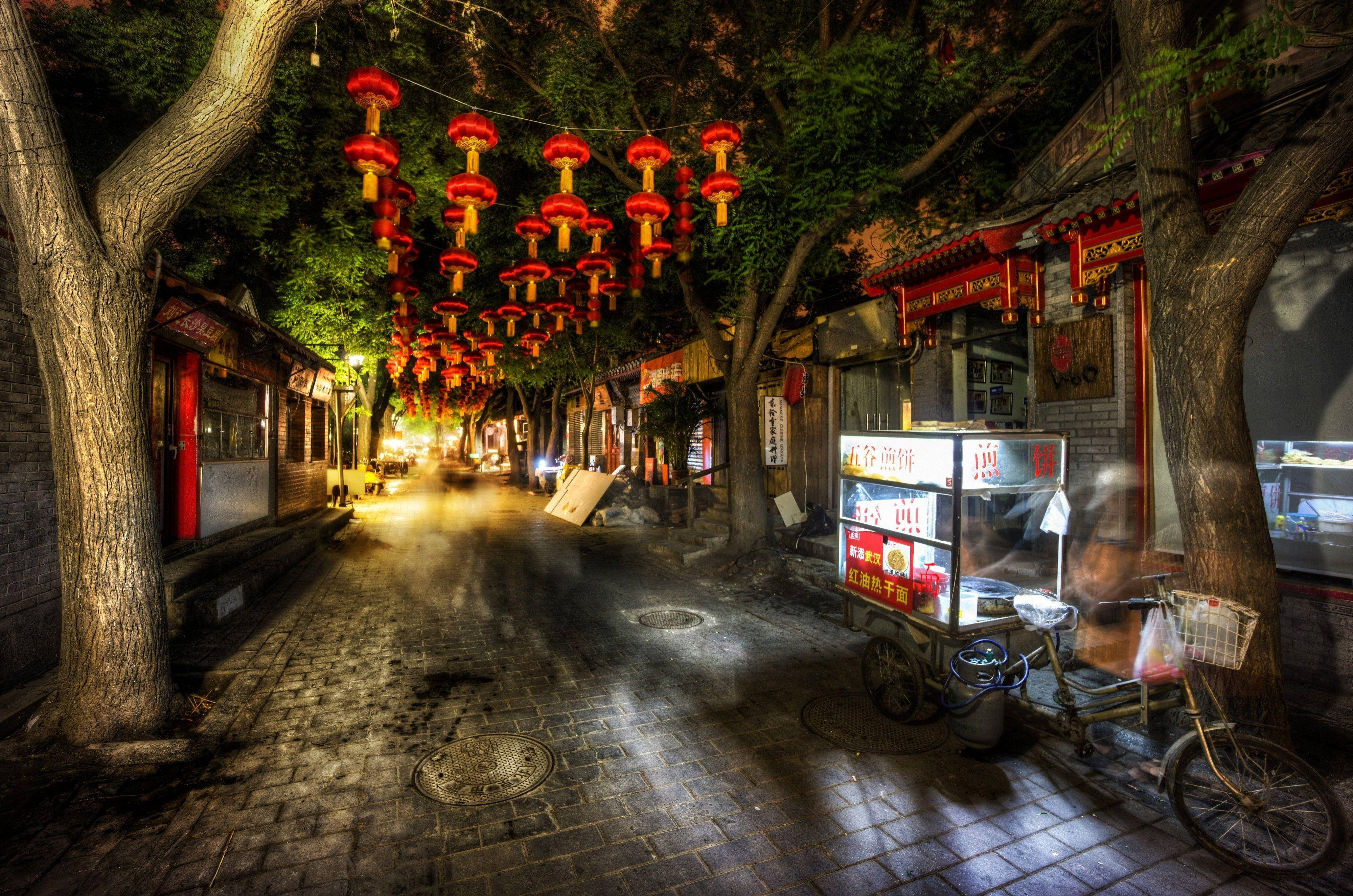 Beijing 桌布 壁紙 — 夜晚一條狹窄的鵝卵石街道，被懸掛在樹木和建築物之間的發光紅色燈籠串照亮。