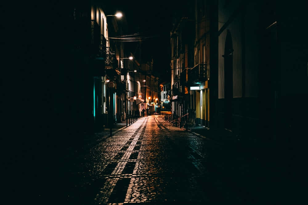 Dark Crossroad 桌布 壁紙 — 夜晚一條狹窄的鵝卵石街道，被溫暖的路燈照亮，在高樓之間投下長長的影子。