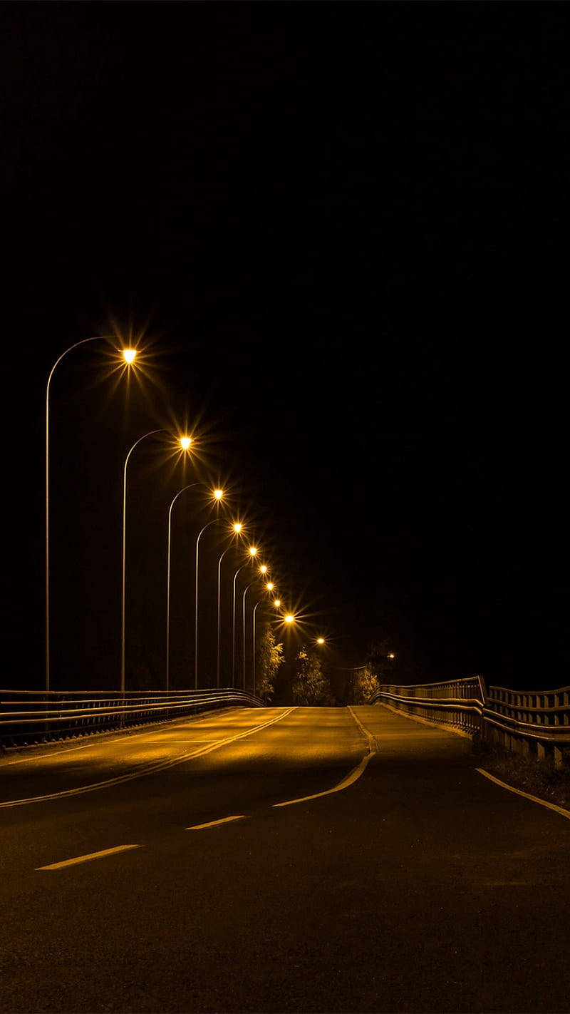 黑夜 桌布 壁紙 — 夜晚一條蜿蜒的道路被一排發光的街燈照亮，將長長的陰影投射到黑暗中。