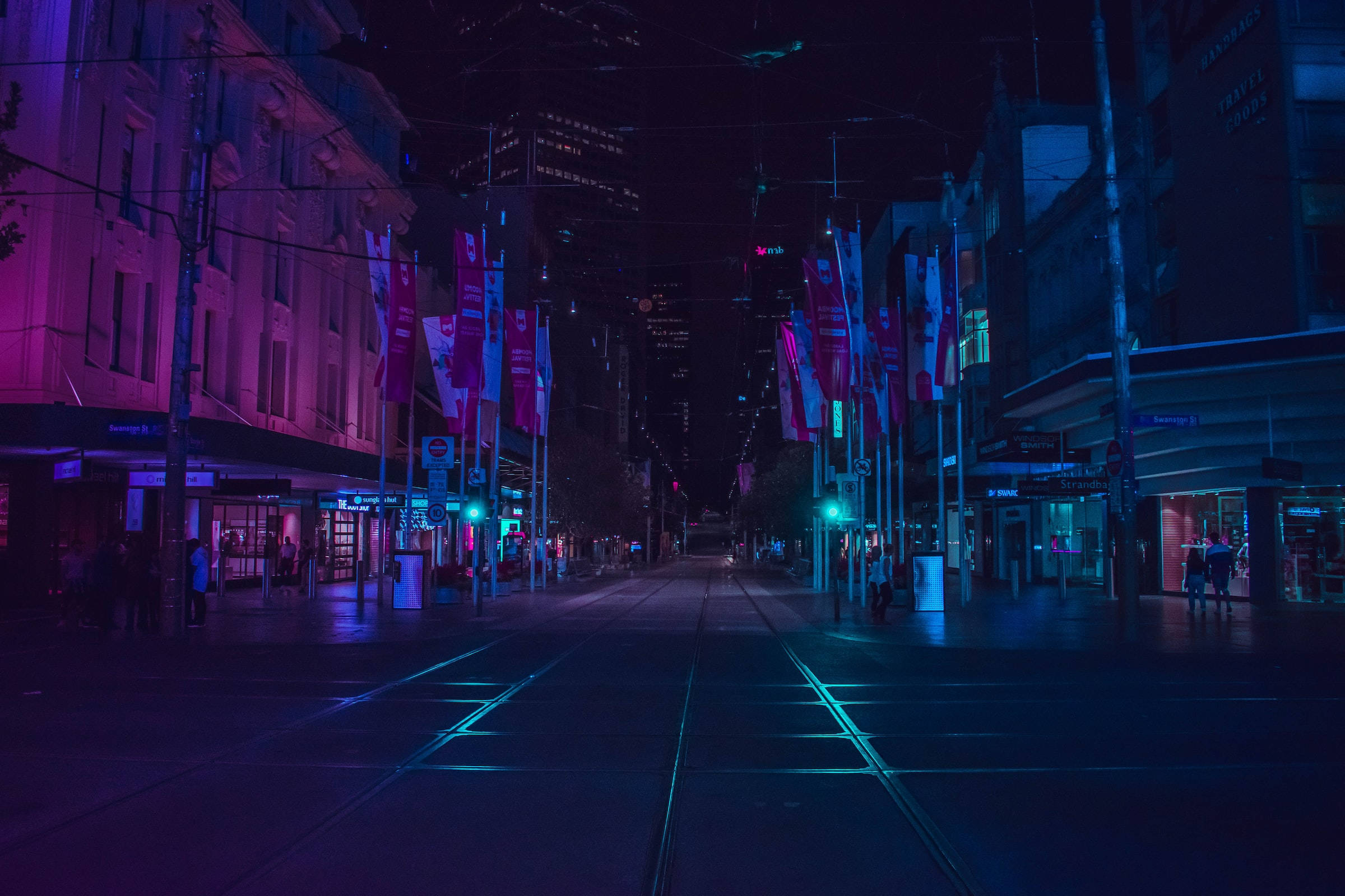 夜空 桌布 壁紙 — 夜晚一條被霓虹燈淹沒的城市街道，紫色和藍色的光芒映照在潮濕的路面和伸向遠方的電車軌道上。