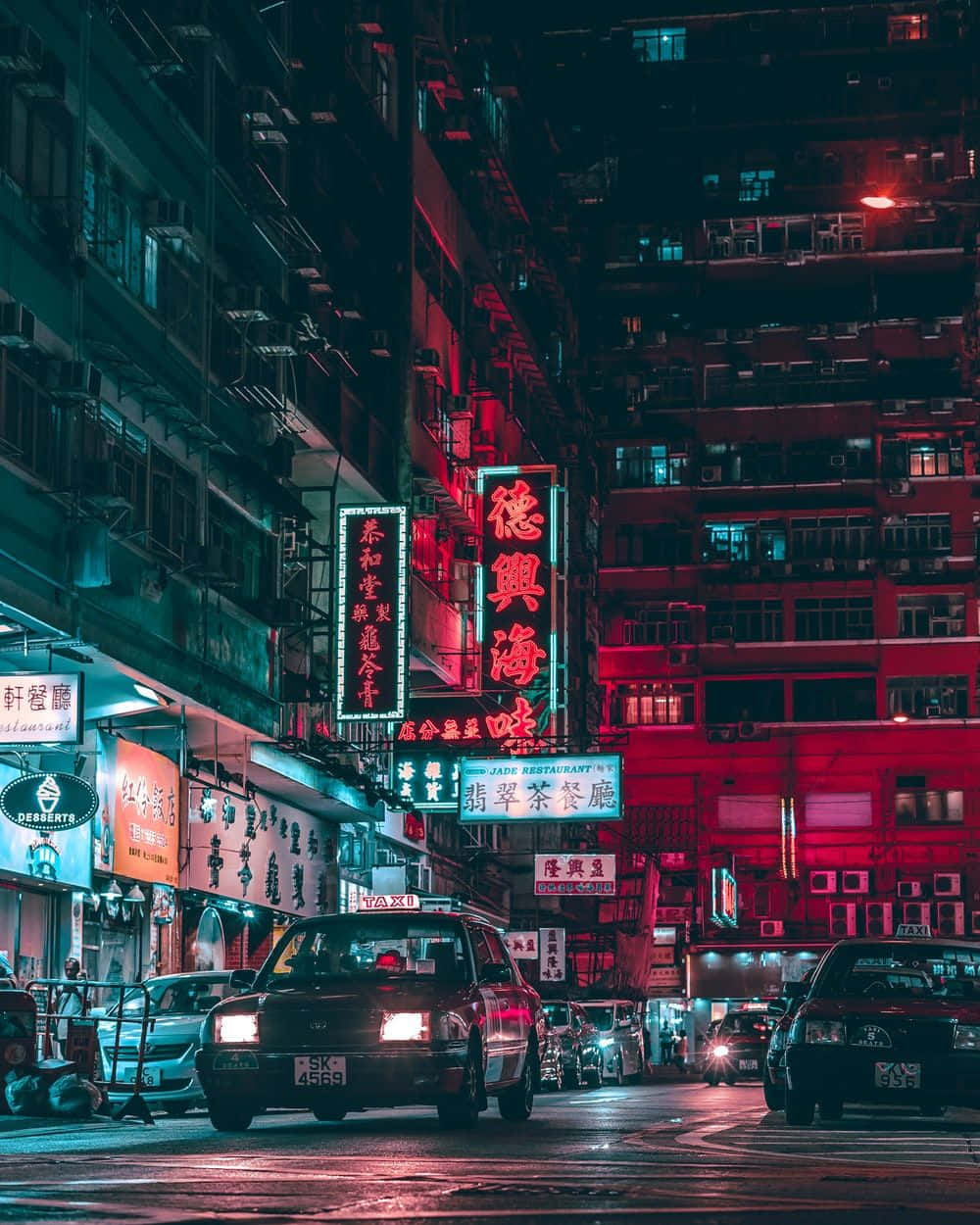 Edgy Aesthetic 桌布 壁紙 — 夜晚一條霓虹燈照亮的城市街道，紅色和藍色的招牌在潮濕的路面上反射，計程車駛過高聳的建築物。
