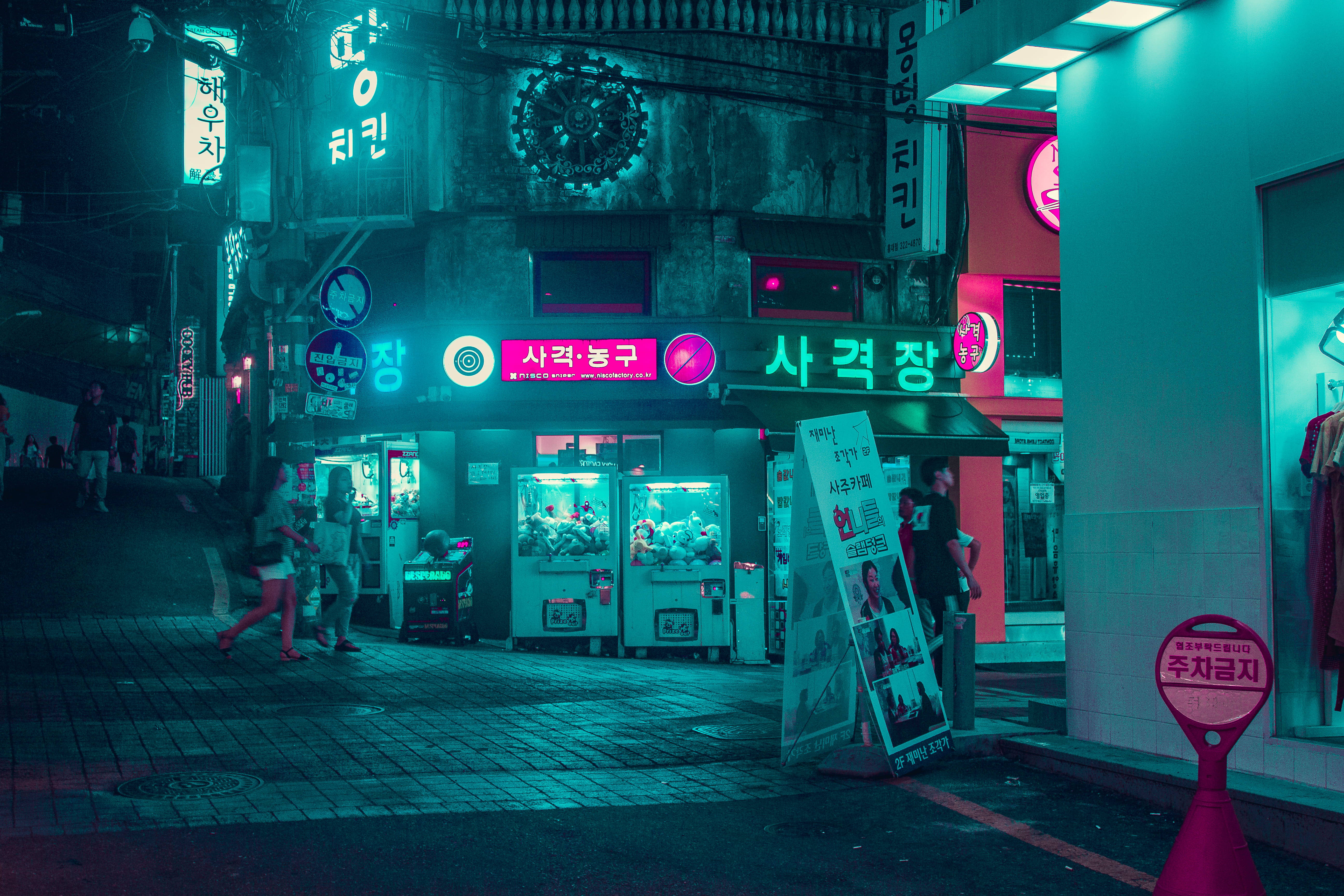 4K Pc 桌布 壁紙 — 夜晚一條霓虹燈照亮的韓國小巷，有發光招牌、夾娃娃機和在濕滑鵝卵石街上行走的行人。