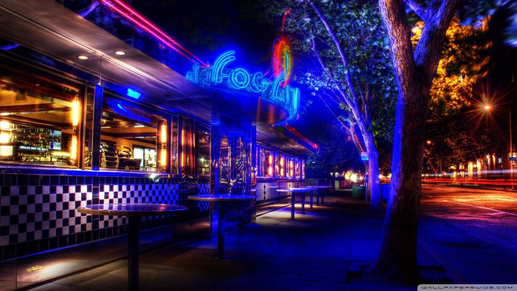 Diner 桌布 壁紙 — 夜晚一間充滿活力的霓虹燈餐廳，藍色和粉紅色的燈光在潮濕的人行道和格子牆上反射。