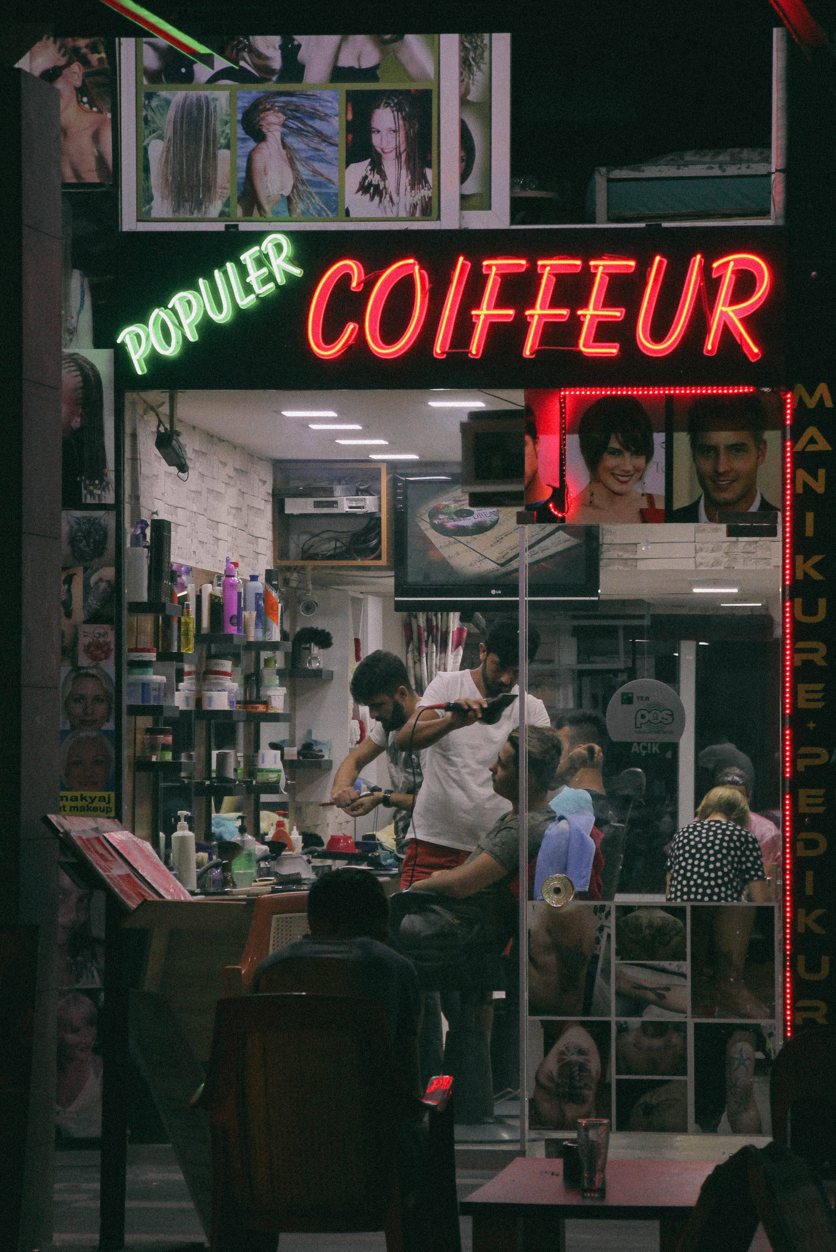 Haircut 桌布 壁紙 — 夜晚一間充滿活力的霓虹燈髮廊，繁忙的市區店面內，造型師正在為顧客服務。