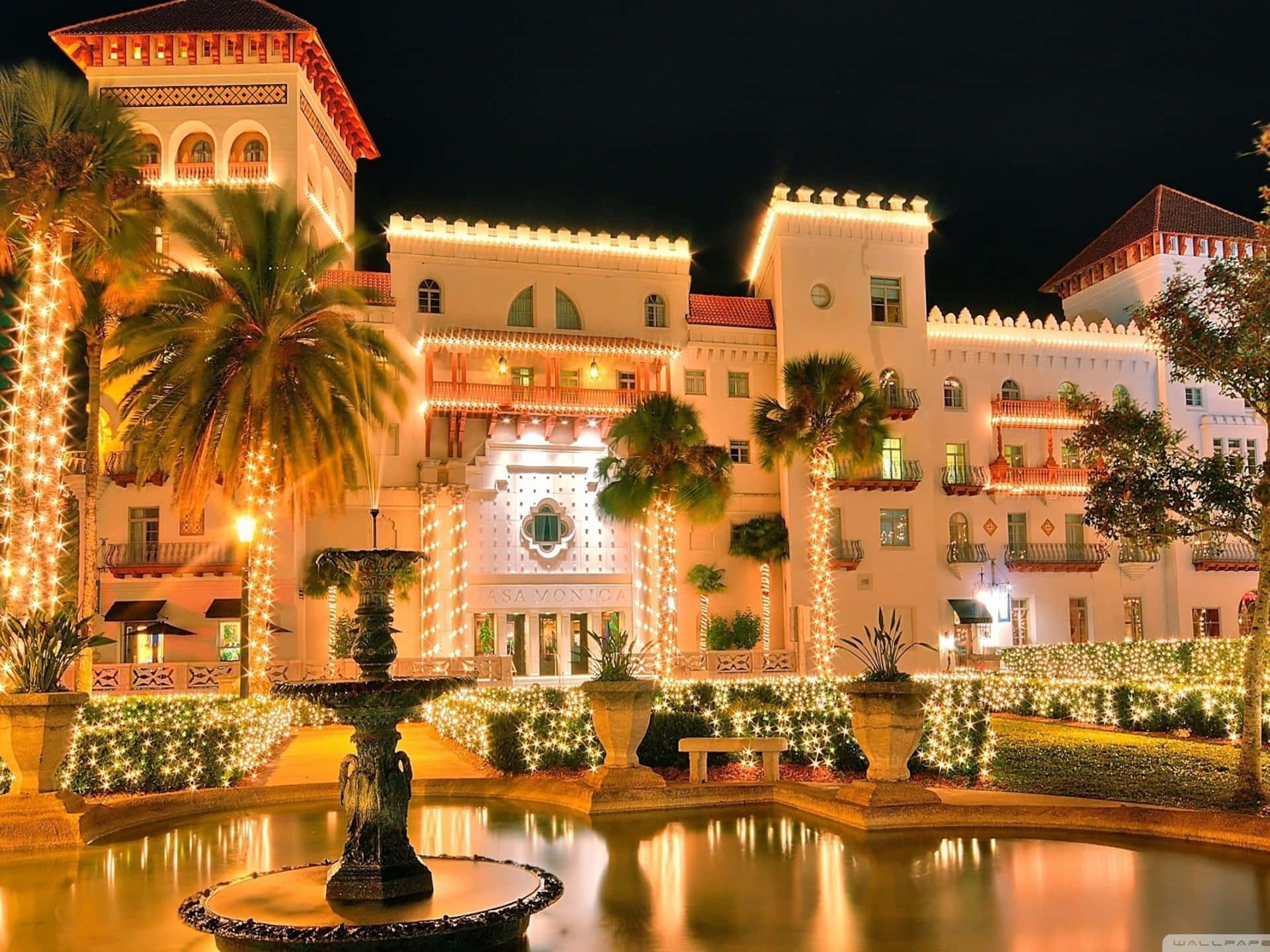 Resort 桌布 壁紙 — 夜晚一間宏偉的西班牙風格酒店，燈火輝煌，棕櫚樹和噴泉反射著金色光芒。