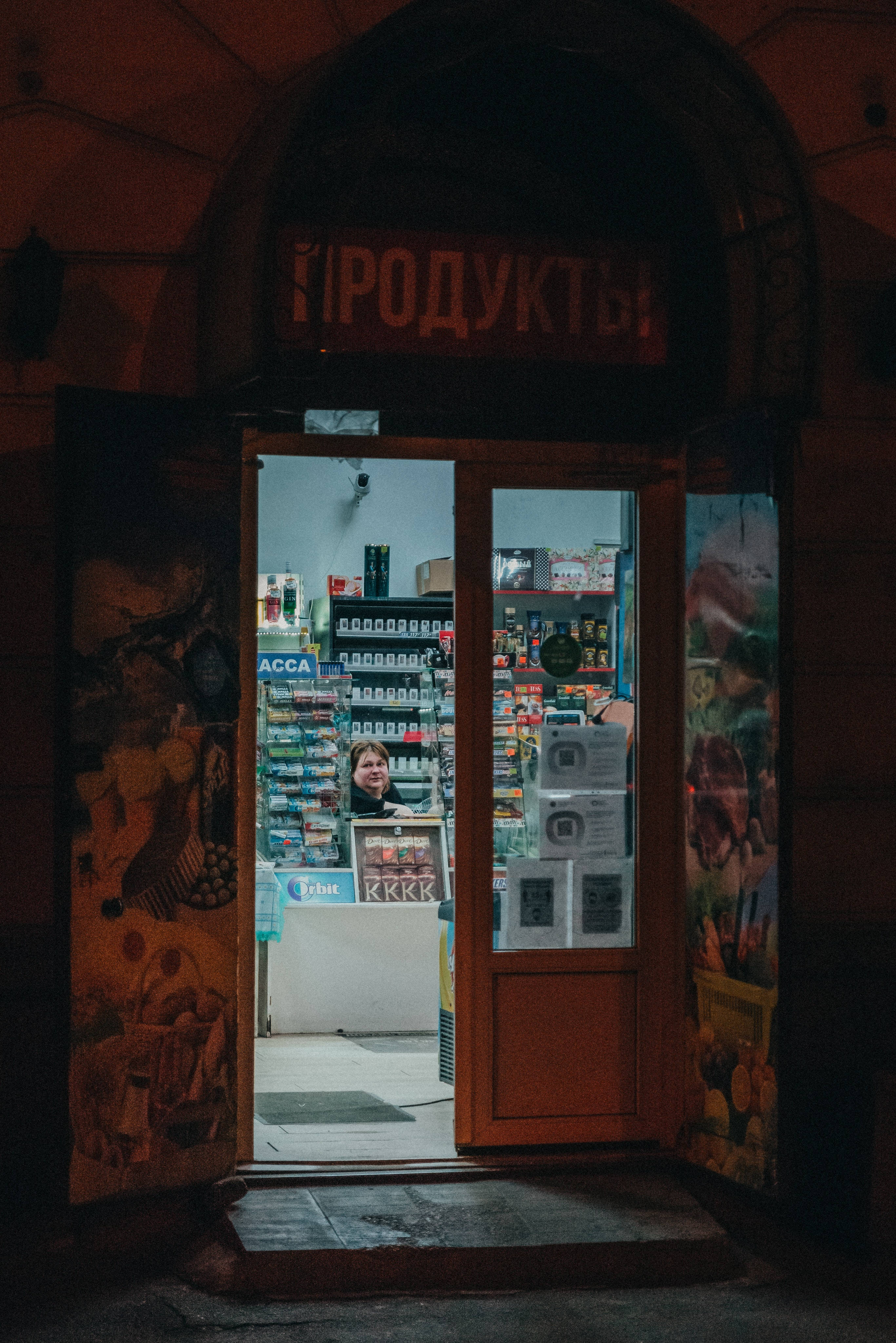 Door 桌布 壁紙 — 夜晚一間溫暖照明的雜貨店門口，露出櫃檯後微笑的女子，周圍是佈滿塗鴉的牆壁和一個寫著「ПРОДУКТЫ」的紅色招牌。