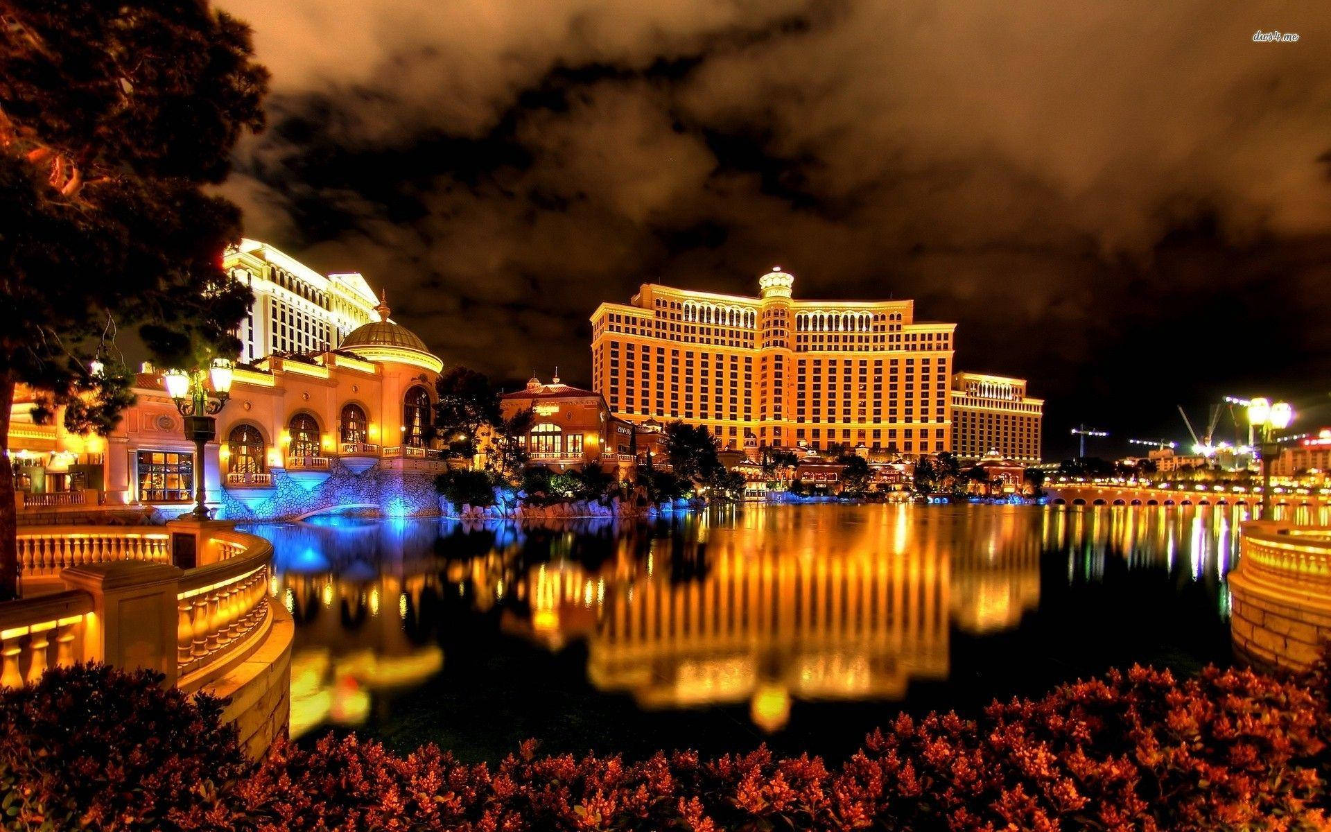 Casino 桌布 壁紙 — 夜晚中，宏偉的照明酒店度假村，金色燈光在平靜的水面上反射，天空多雲。