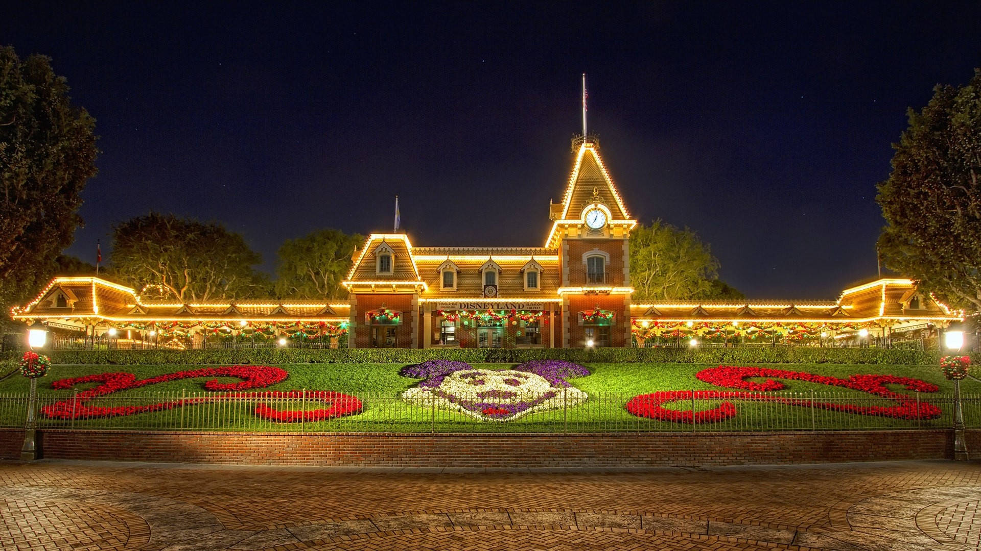 Disneyland Desktop 桌布 壁紙 — 夜晚中一座宏偉的照明車站建築，裝飾著節日燈飾和一個用紅色和紫色花朵拼出「2023」的鮮豔花卉展示。
