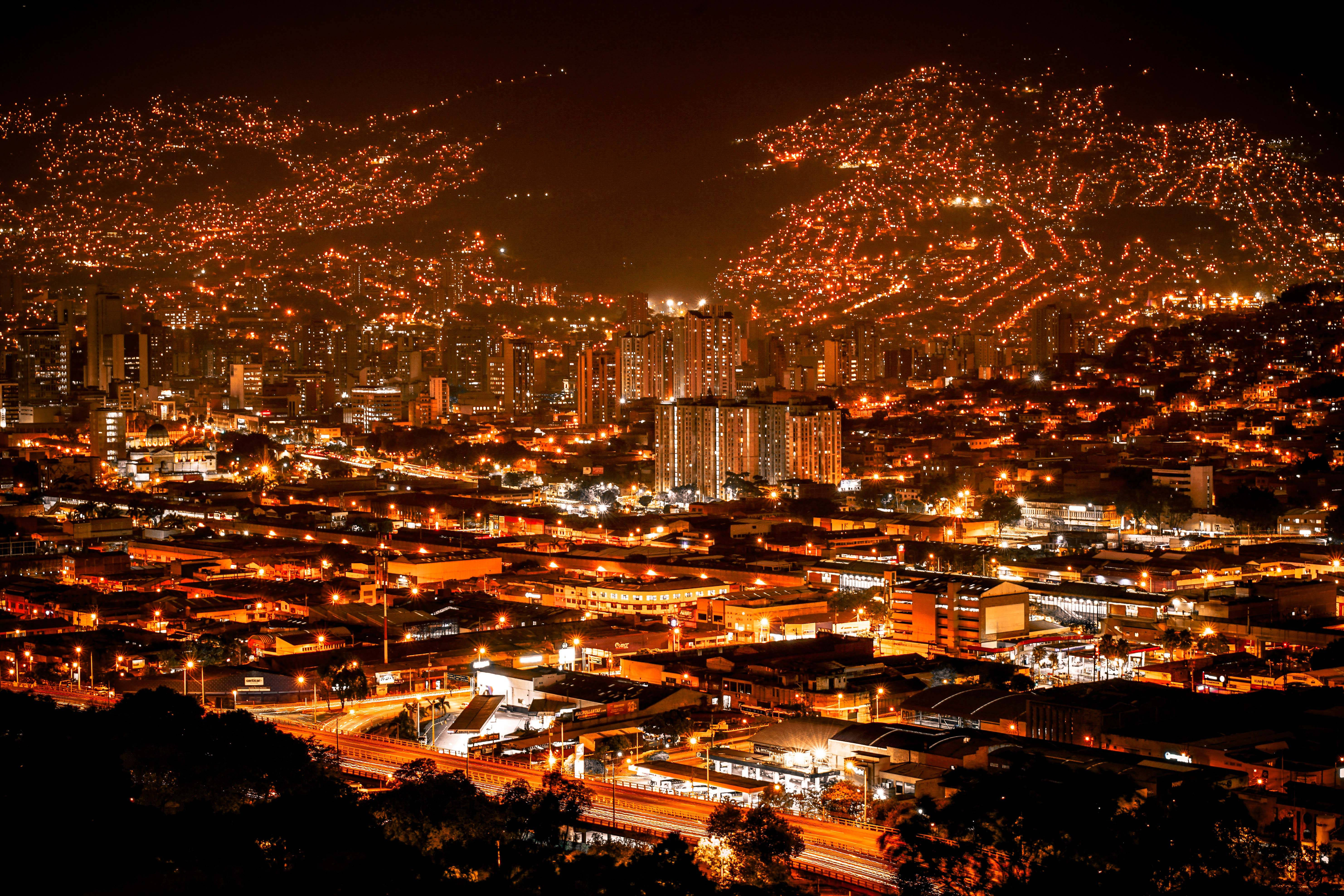 Medellin 桌布 壁紙 — 夜晚中一座廣闊的城市，溫暖的橙色燈光照亮密集的城市擴展區和覆蓋著發光房屋的山坡。