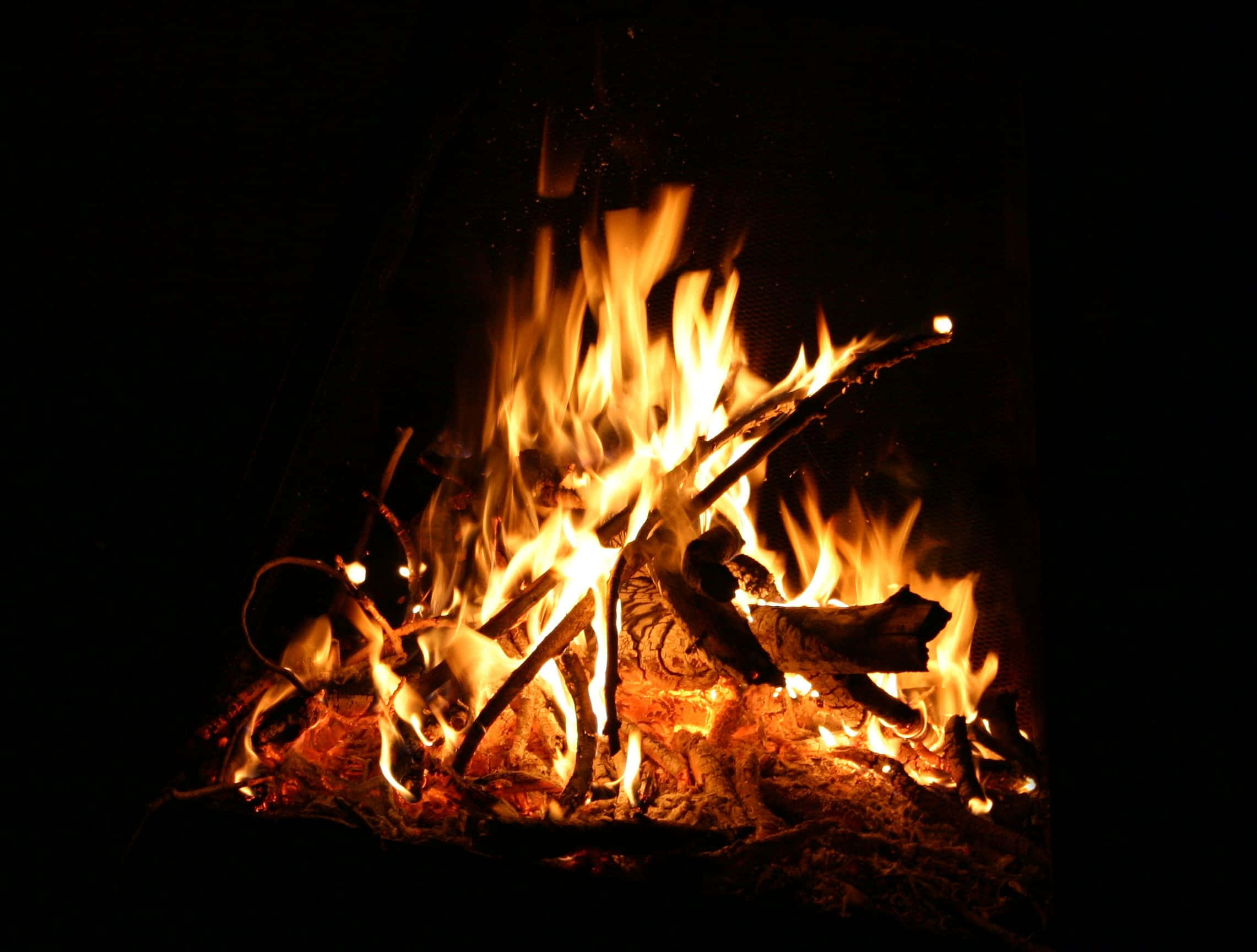 Campfire 桌布 壁紙 — 夜晚中熊熊燃燒的營火，帶有明亮的橙色火焰和發光的餘燼，背景為黑色。