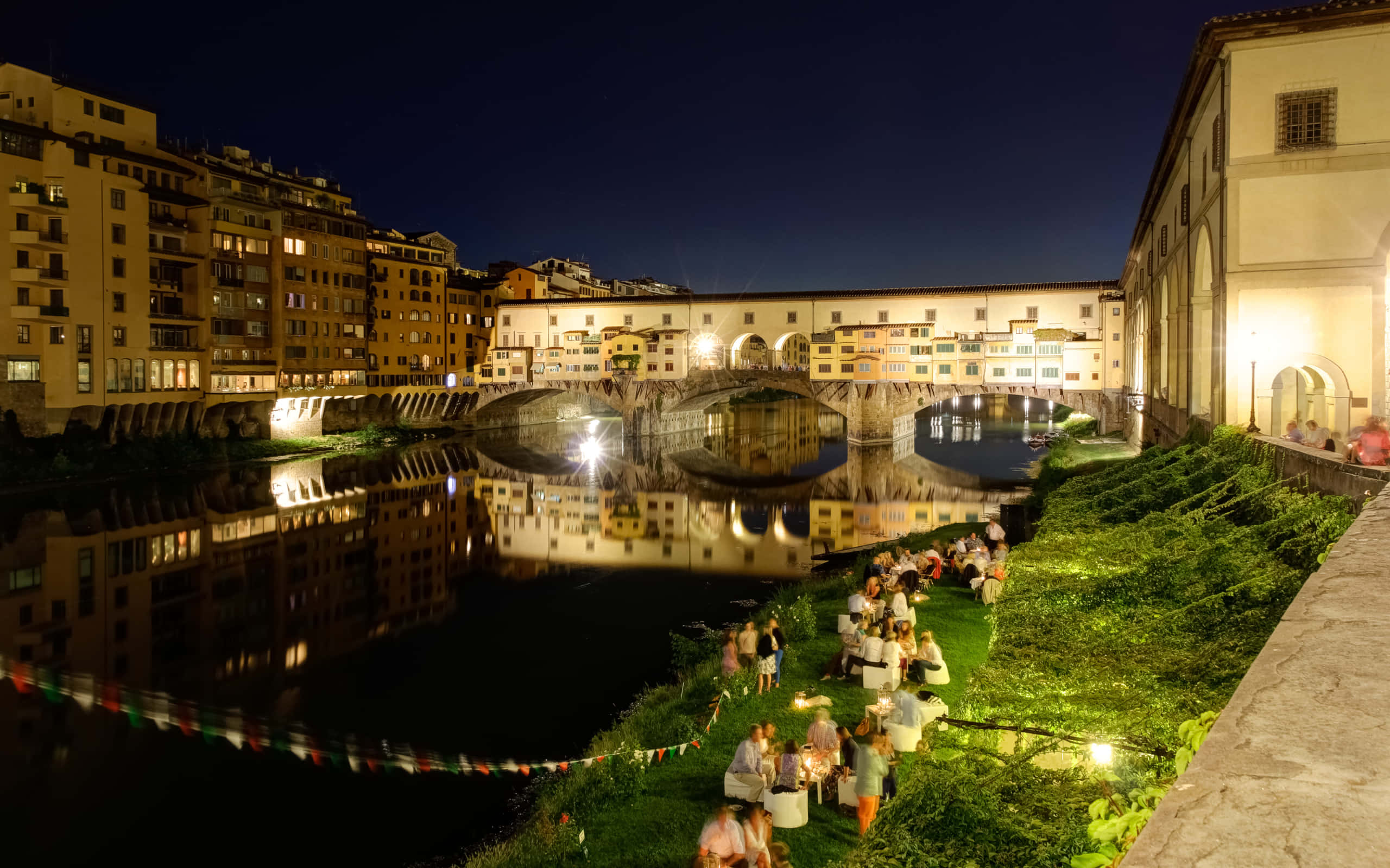 City View 桌布 壁紙 — 夜晚亮燈的 Ponte Vecchio 橋，人們在溫暖燈光下的草河岸用餐。