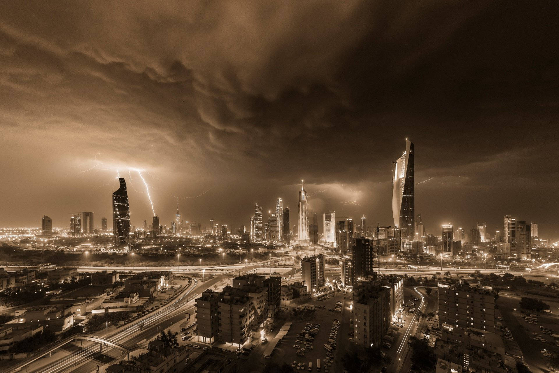 Kuwait 桌布 壁紙 — 夜晚以深褐色調呈現的戲劇性城市景觀，閃電劃過高聳的摩天大樓，下方街道閃爍著光芒。