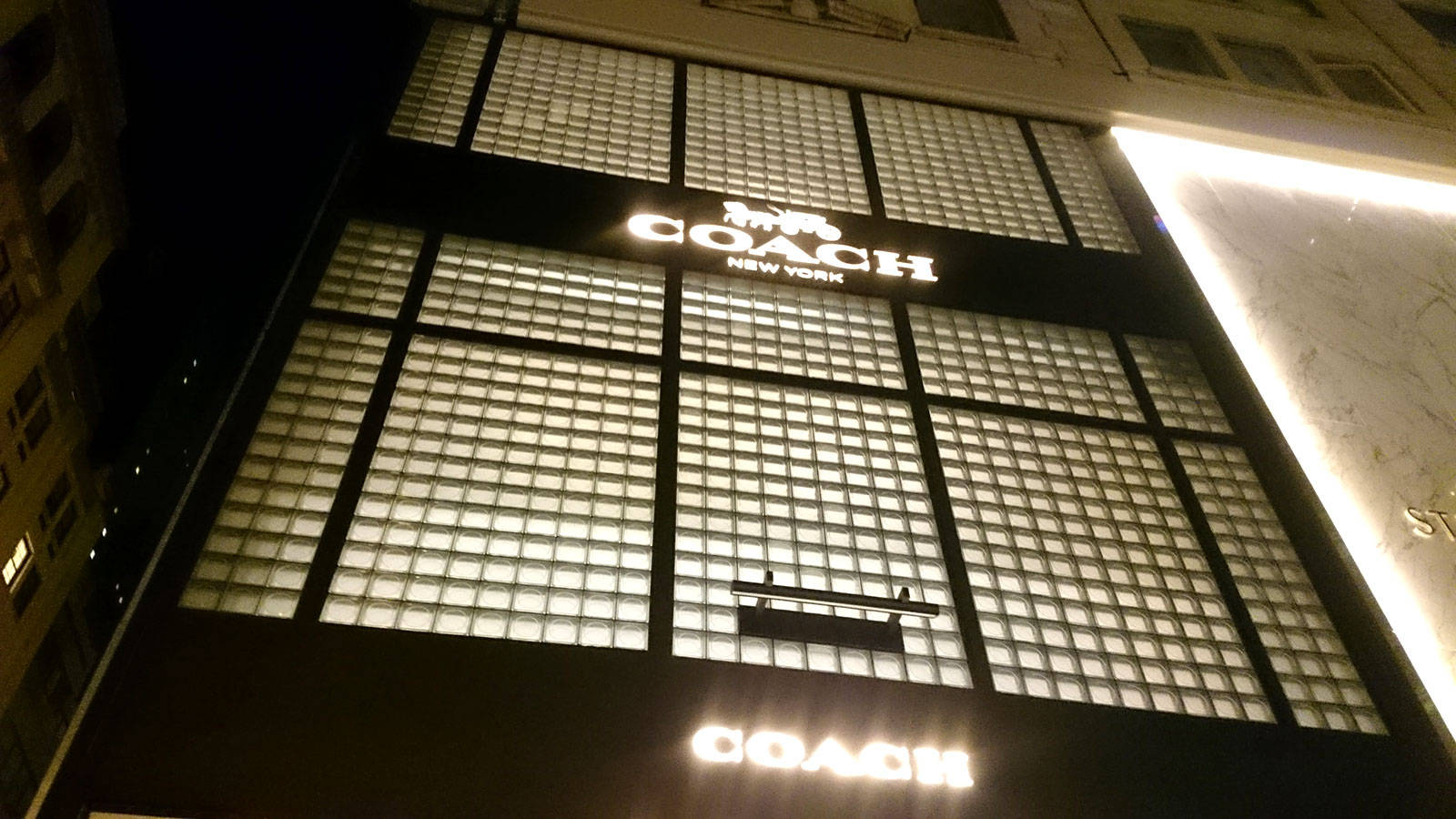 Coach 桌布 壁紙 — 夜晚低角度拍攝的 Coach New York 商店外牆，帶有發光的玻璃磚窗和照明招牌。