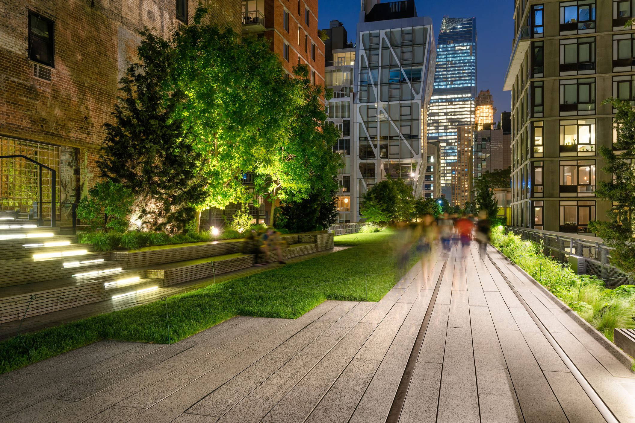 New York 桌布 壁紙 — 夜晚充滿活力的城市公園，發光的綠色樹木和模糊的行人在被照亮的摩天大樓環繞的木質小徑上。