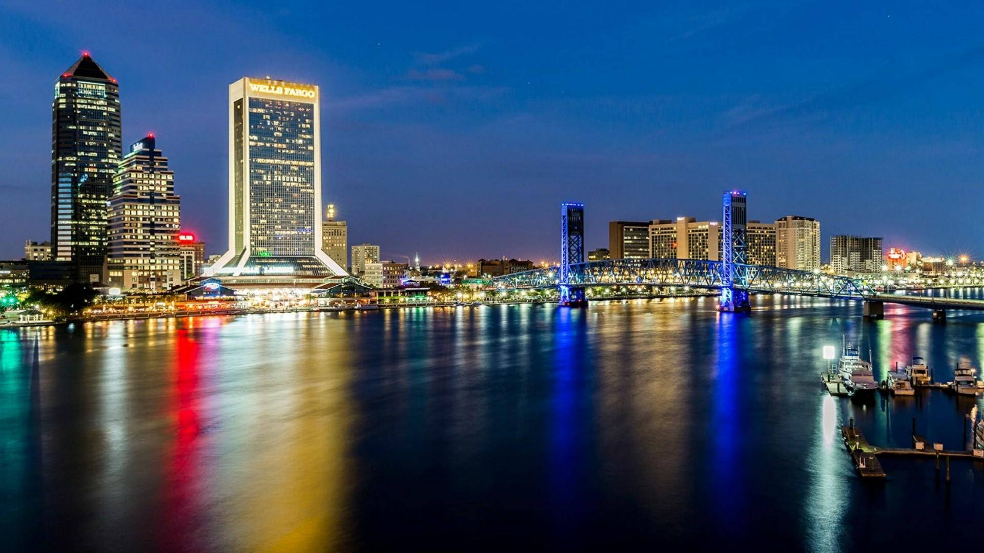 Jacksonville 桌布 壁紙 — 夜晚充滿活力的城市天際線，燈火通明的摩天大樓和發光的橋樑倒映在平靜的水面上。