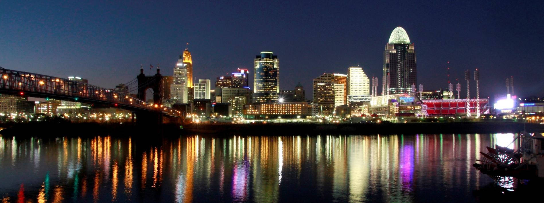 Cincinnati 桌布 壁紙 — 夜晚充滿活力的城市天際線，燈火通明的摩天大樓和橋樑倒映在平靜的水面上。
