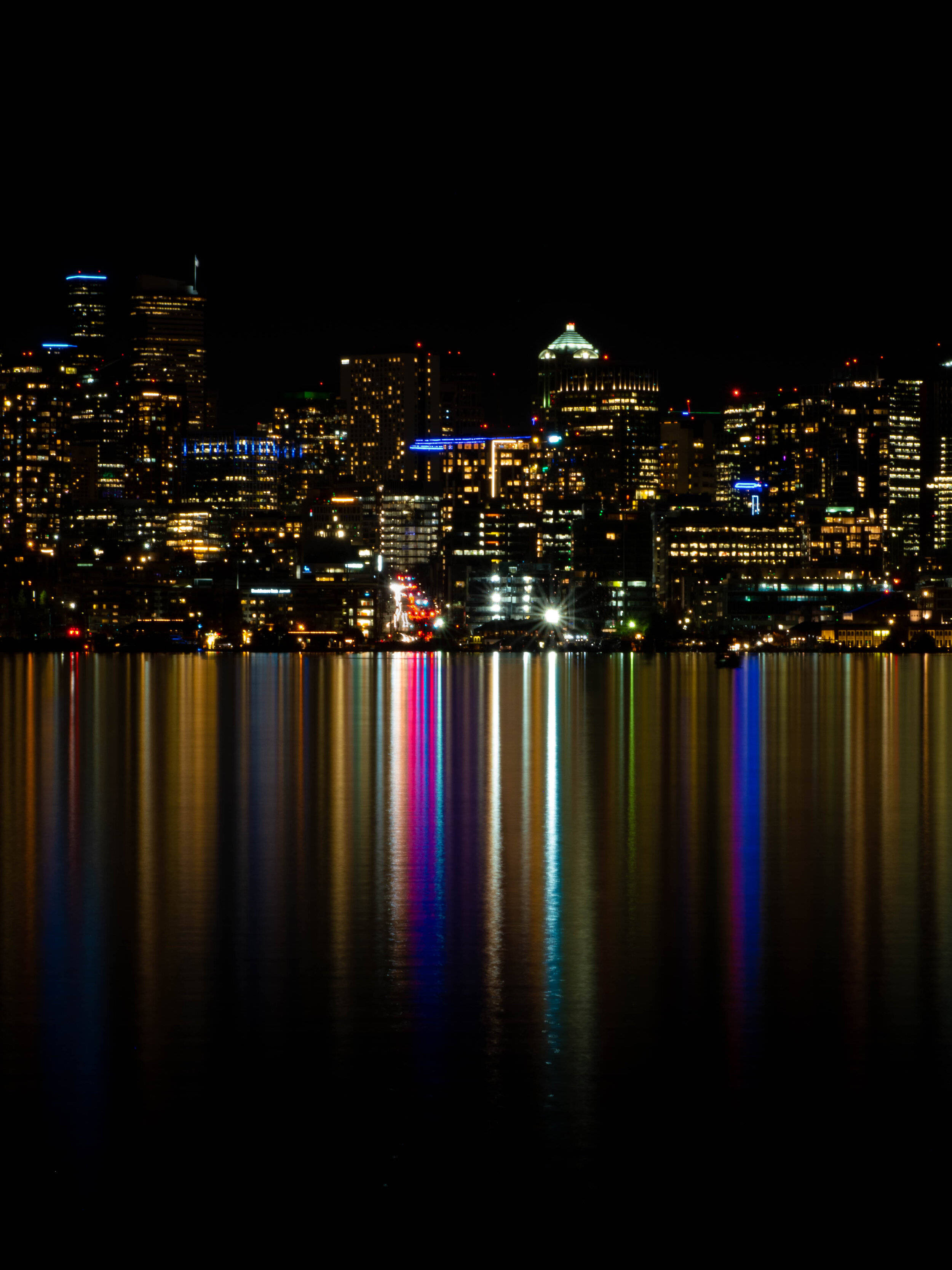 Seattle Skyline 桌布 壁紙 — 夜晚充滿活力的城市天際線倒映在平靜的水面上，色彩繽紛的燈光形成閃爍的垂直條紋。