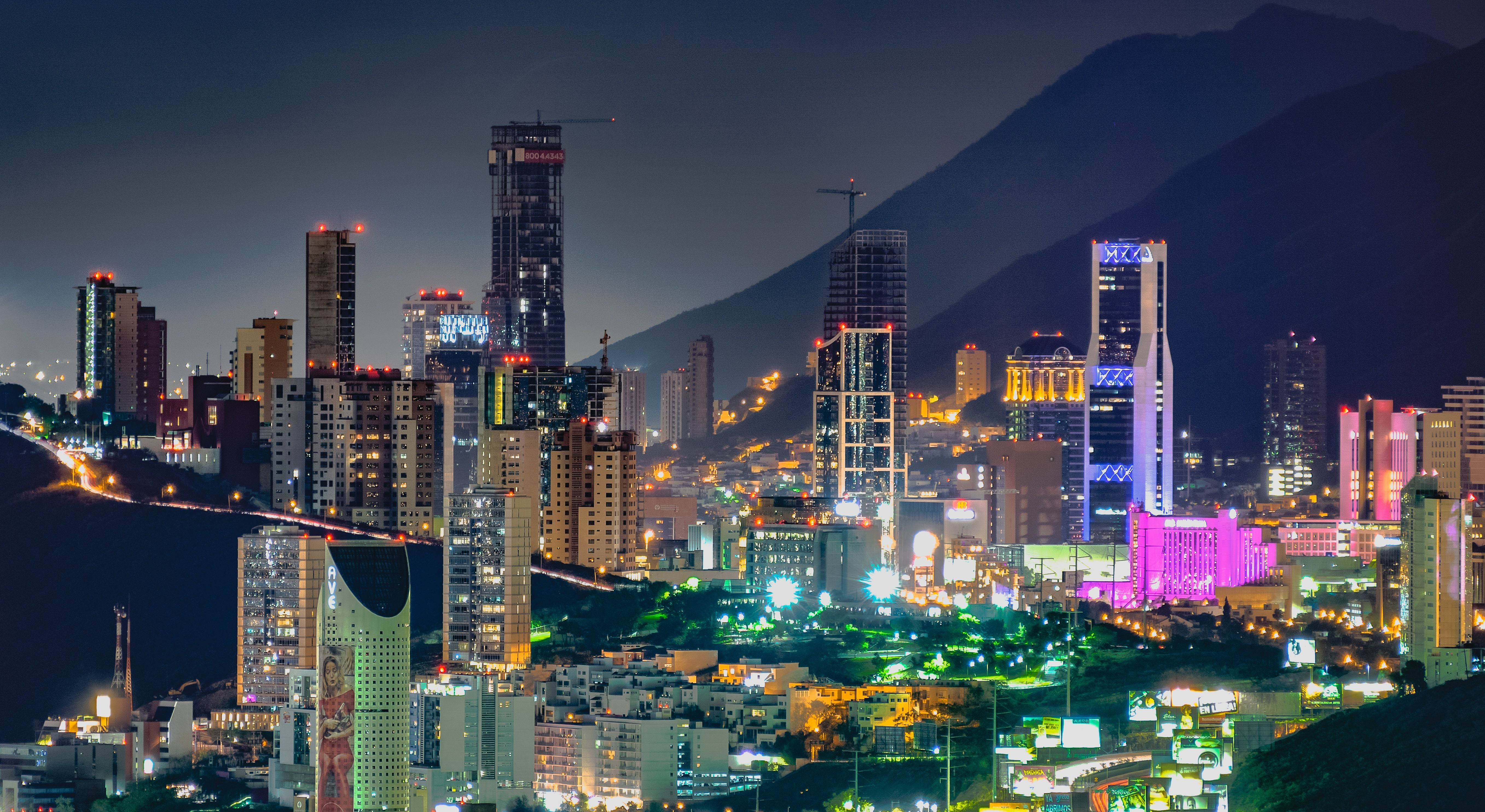 Monterrey 桌布 壁紙 — 夜晚充滿活力的城市景觀，有燈火通明的摩天大樓和背景中的山脈。