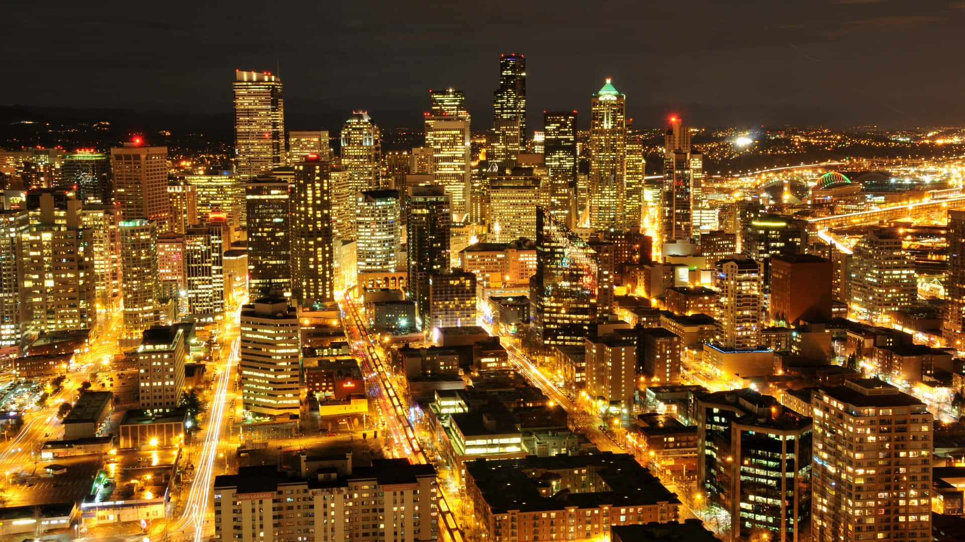 City At Night 桌布 壁紙 — 夜晚充滿活力的城市景觀鳥瞰圖，閃爍的摩天大樓和劃過的街燈照亮了城市廣闊的區域。