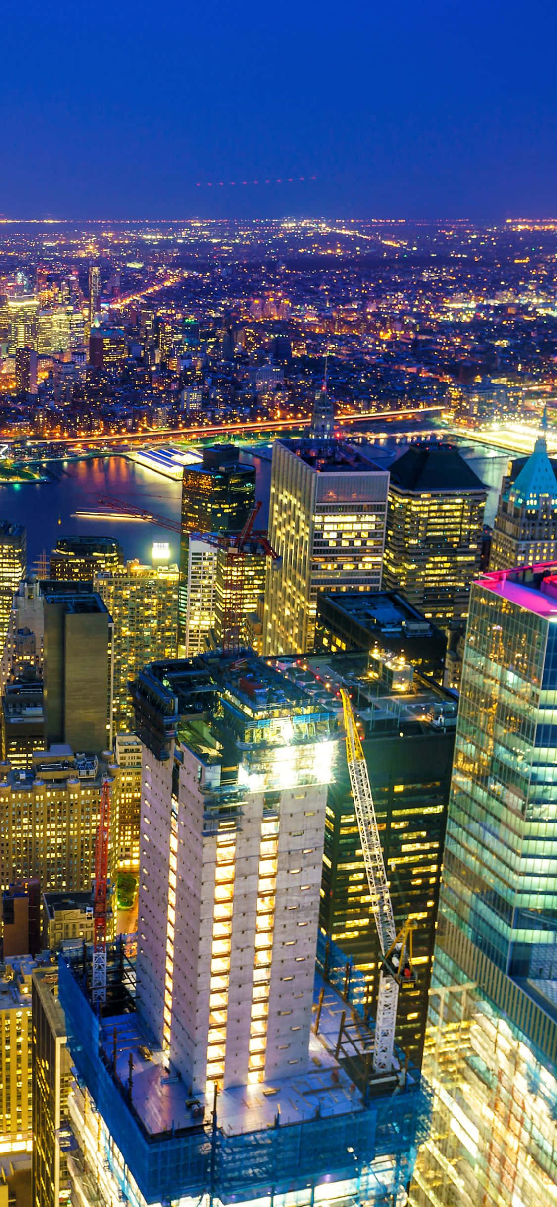 iPhone X New York 桌布 壁紙 — 夜晚充滿活力的城市景觀鳥瞰圖，閃爍的摩天大樓和反射燈光的河流。