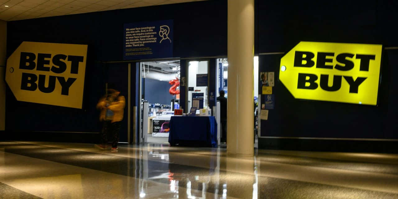 Best Buy 桌布 壁紙 — 夜晚光線昏暗的Best Buy商店入口，帶有發光的黃色標誌和一個模糊的行人走過。
