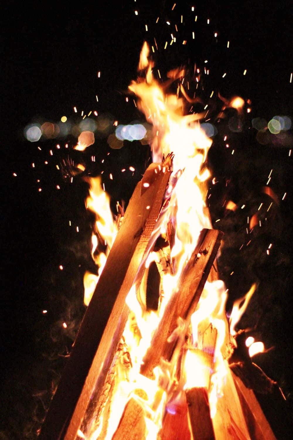 Beach Bonfire 桌布 壁紙 — 夜晚咆哮的營火，明亮的橙色火焰和飛舞的火星，映襯著昏暗的天空和模糊的城市燈光。