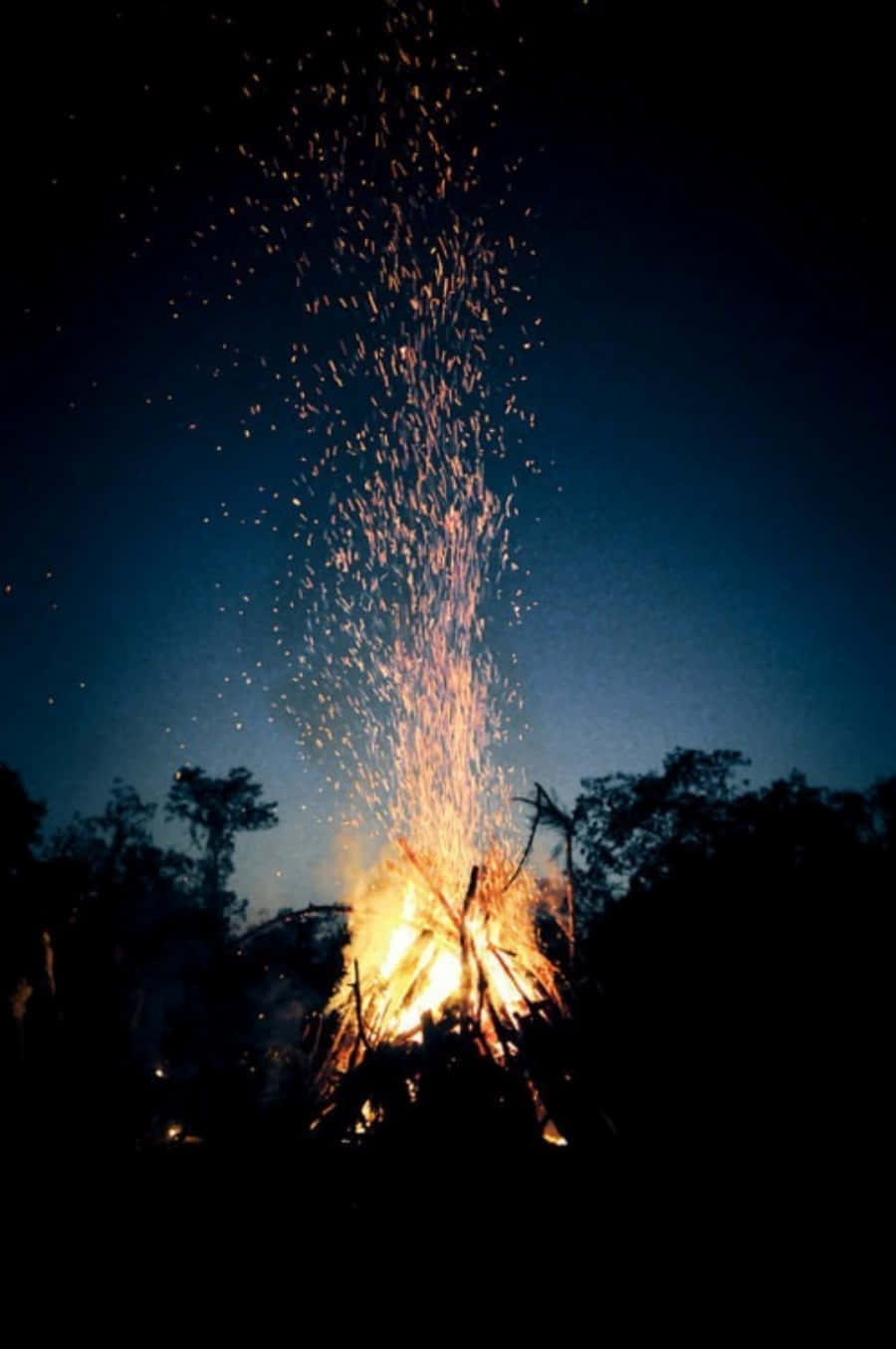 Tumblr 桌布 壁紙 — 夜晚咆哮的營火將金色火花送向深藍色天空，上方是剪影樹木。