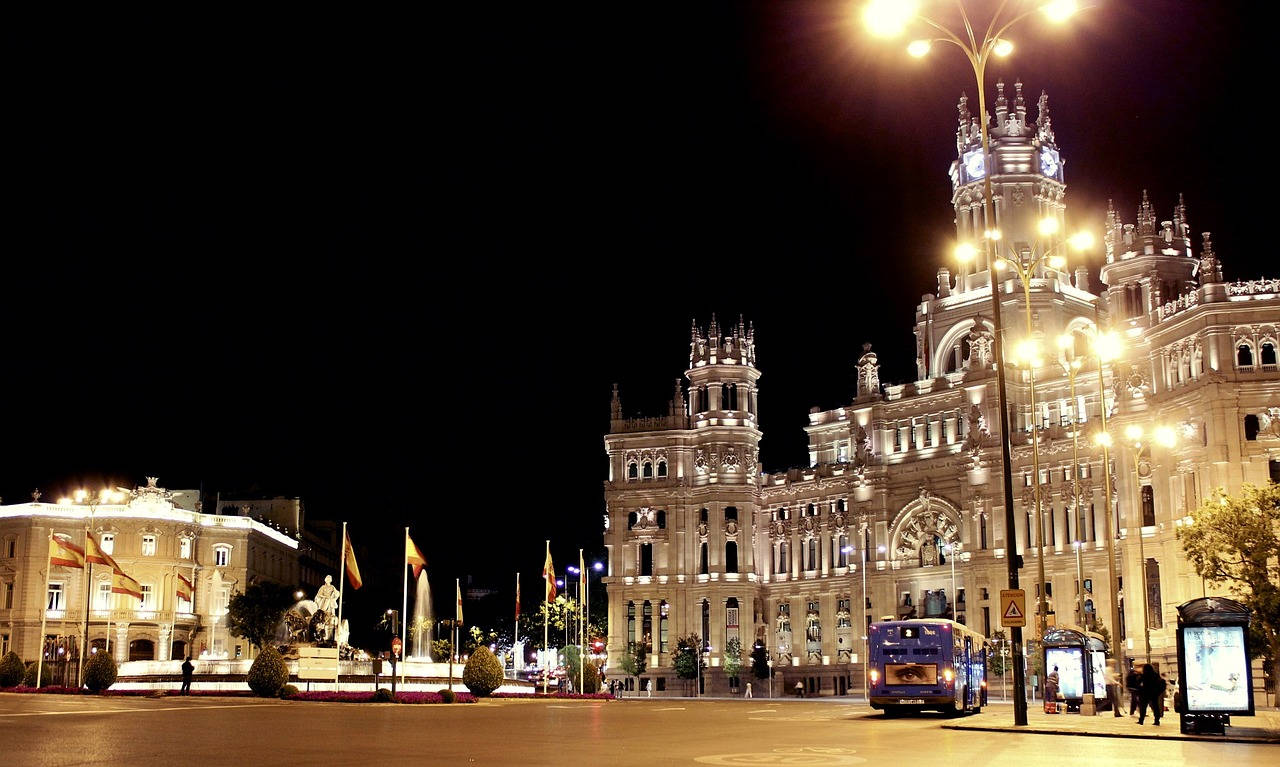 Madrid 桌布 壁紙 — 夜晚在繁華的城市廣場上，宏偉的新古典主義建築被照亮，有路燈和一輛巴士經過。