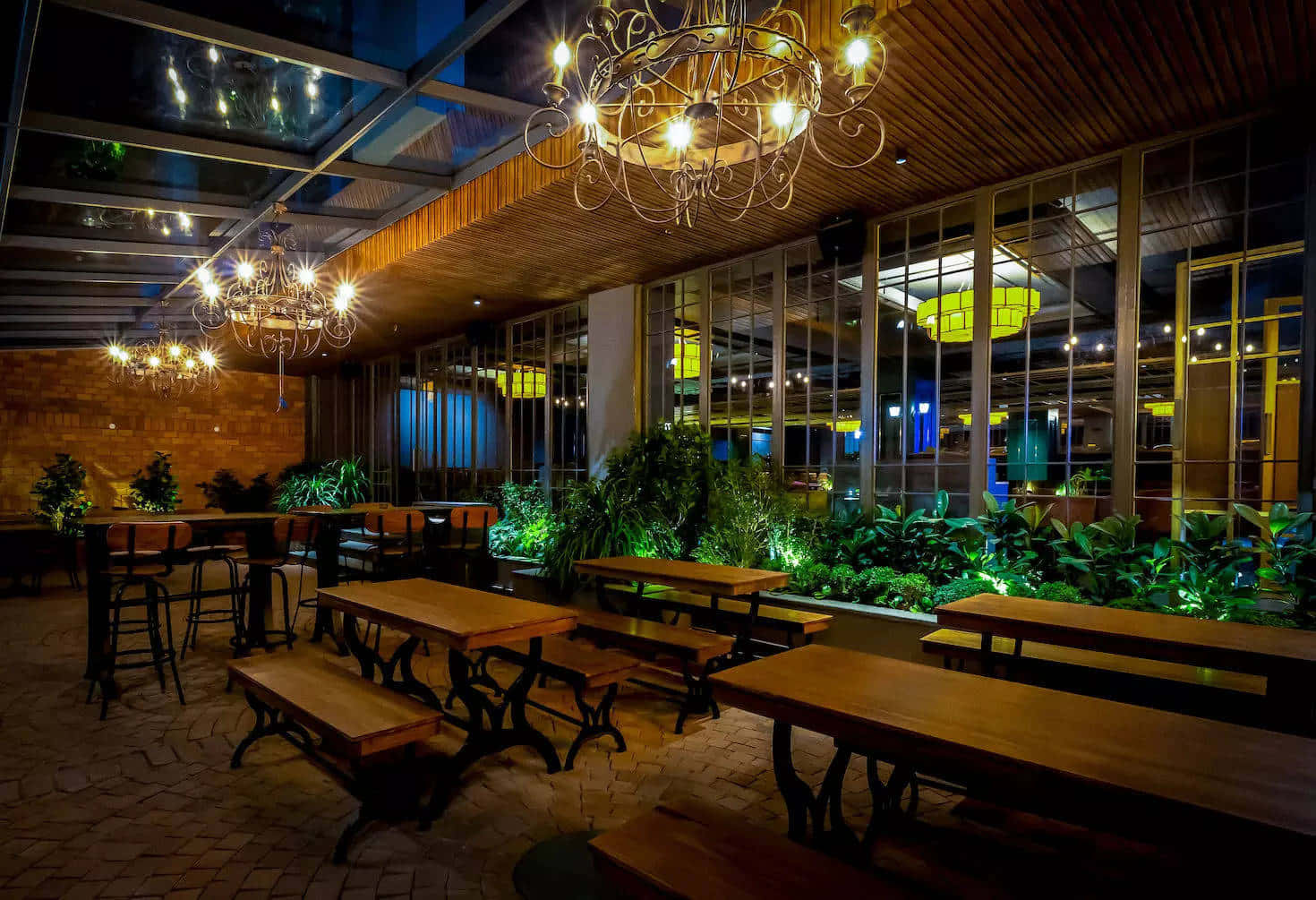 Pub 桌布 壁紙 — 夜晚在華麗吊燈下，溫暖照明的室內用餐區，配有木桌、長凳和茂盛的綠色植物。