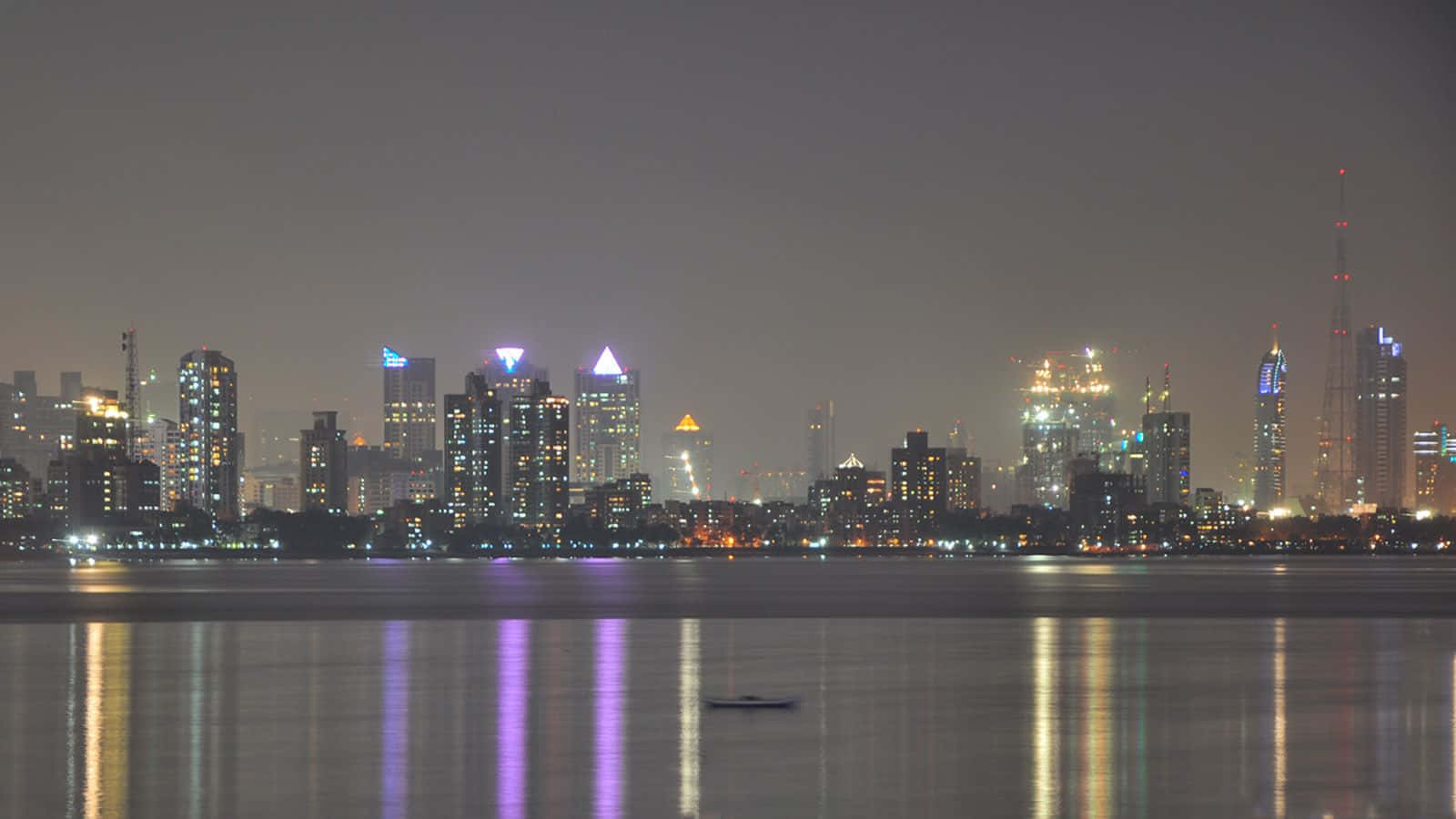 Mumbai 桌布 壁紙 — 夜晚城市天際線，燈火通明的摩天大樓倒映在平靜的水面上，附近漂浮著一艘小船。