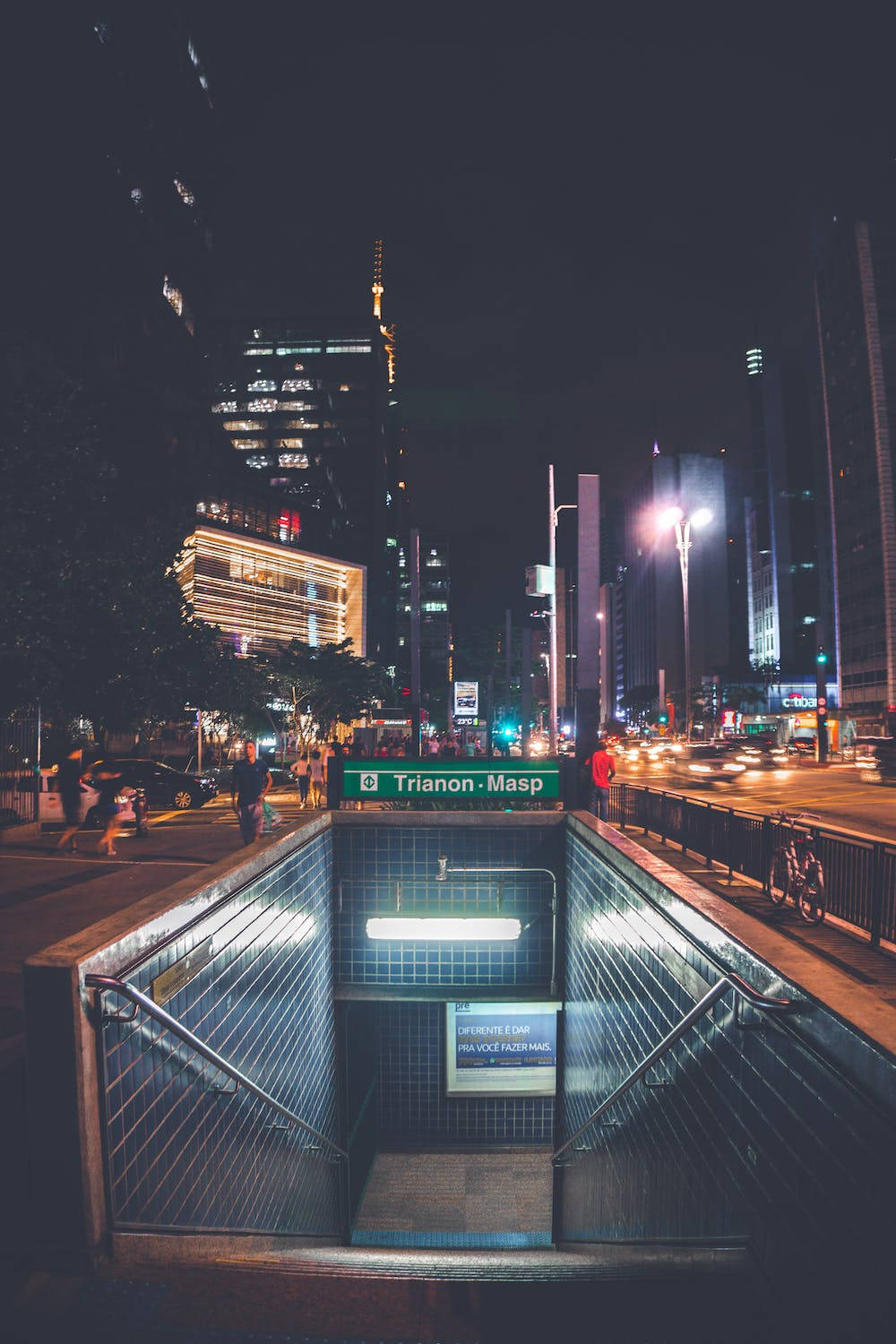 夜景城市 桌布 壁紙 — 夜晚城市景觀，一個燈火通明的地鐵入口，樓梯向下通往瓷磚車站，上方是高聳的摩天大樓和發光的街燈。