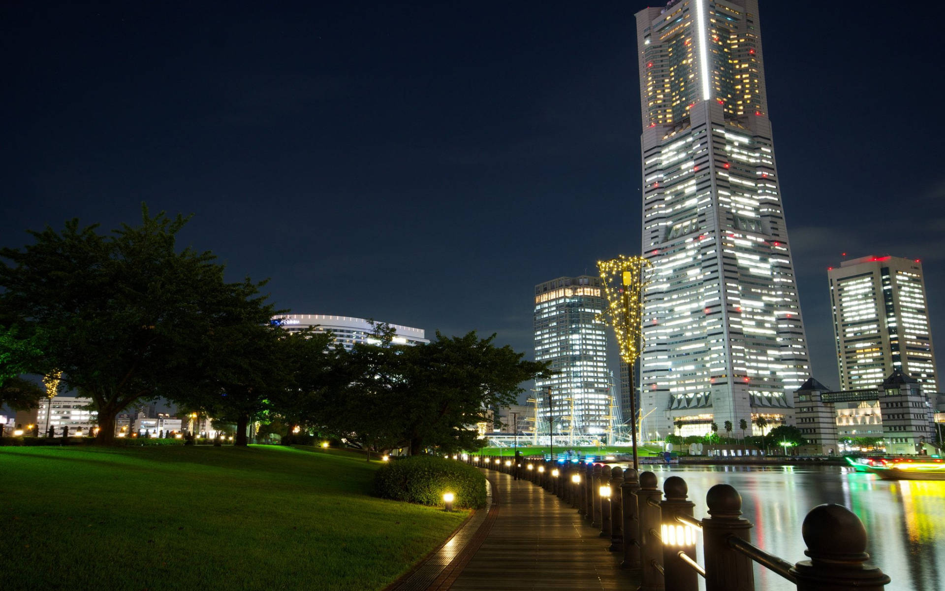 Yokohama 桌布 壁紙 — 夜晚城市景觀，河邊有一座發光的摩天大樓，旁邊有照明步道和綠色公園。