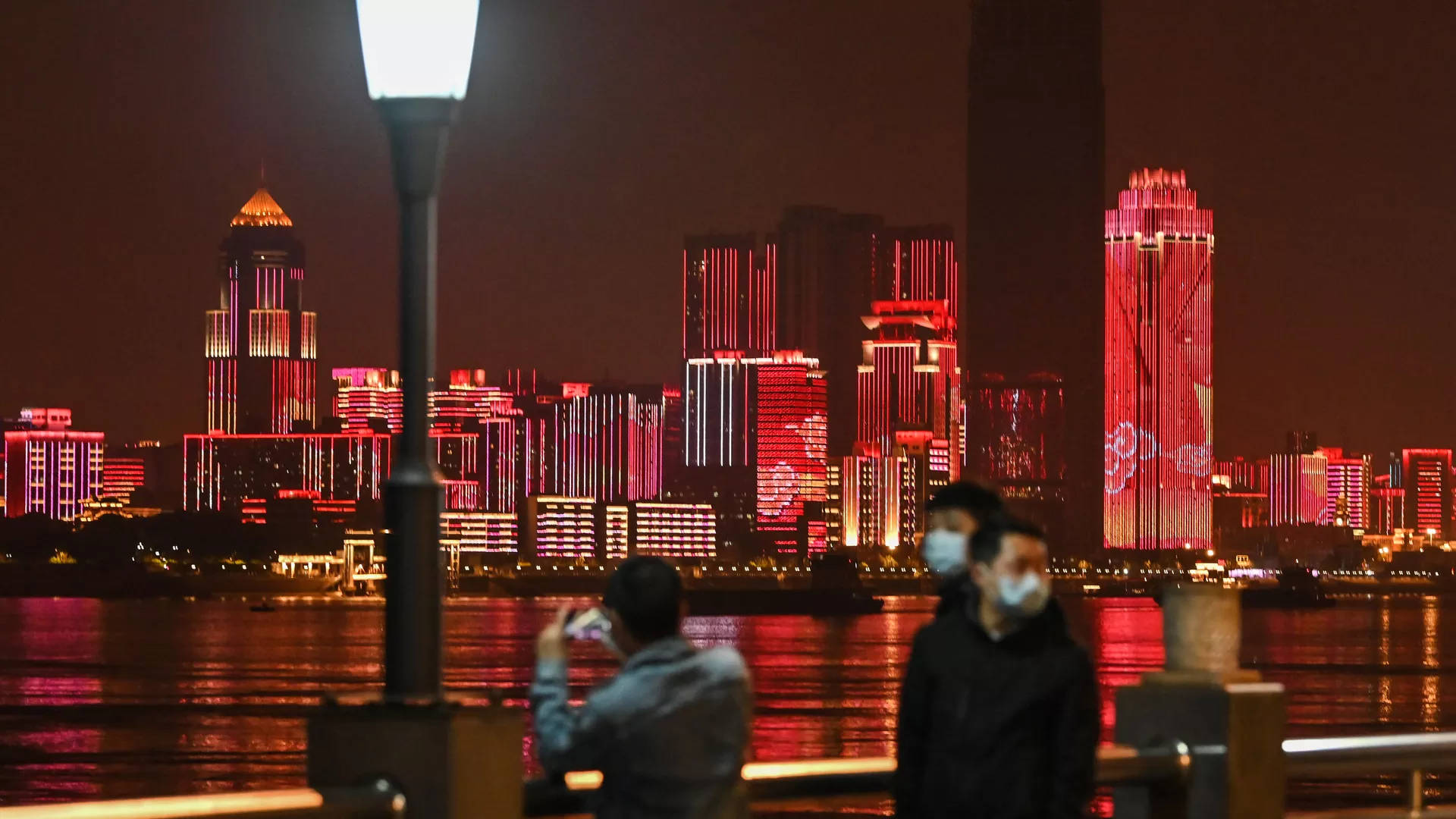 Wuhan 桌布 壁紙 — 夜晚城市景觀，紅色發光的摩天大樓倒映在水中，人們沿著河濱長廊散步。