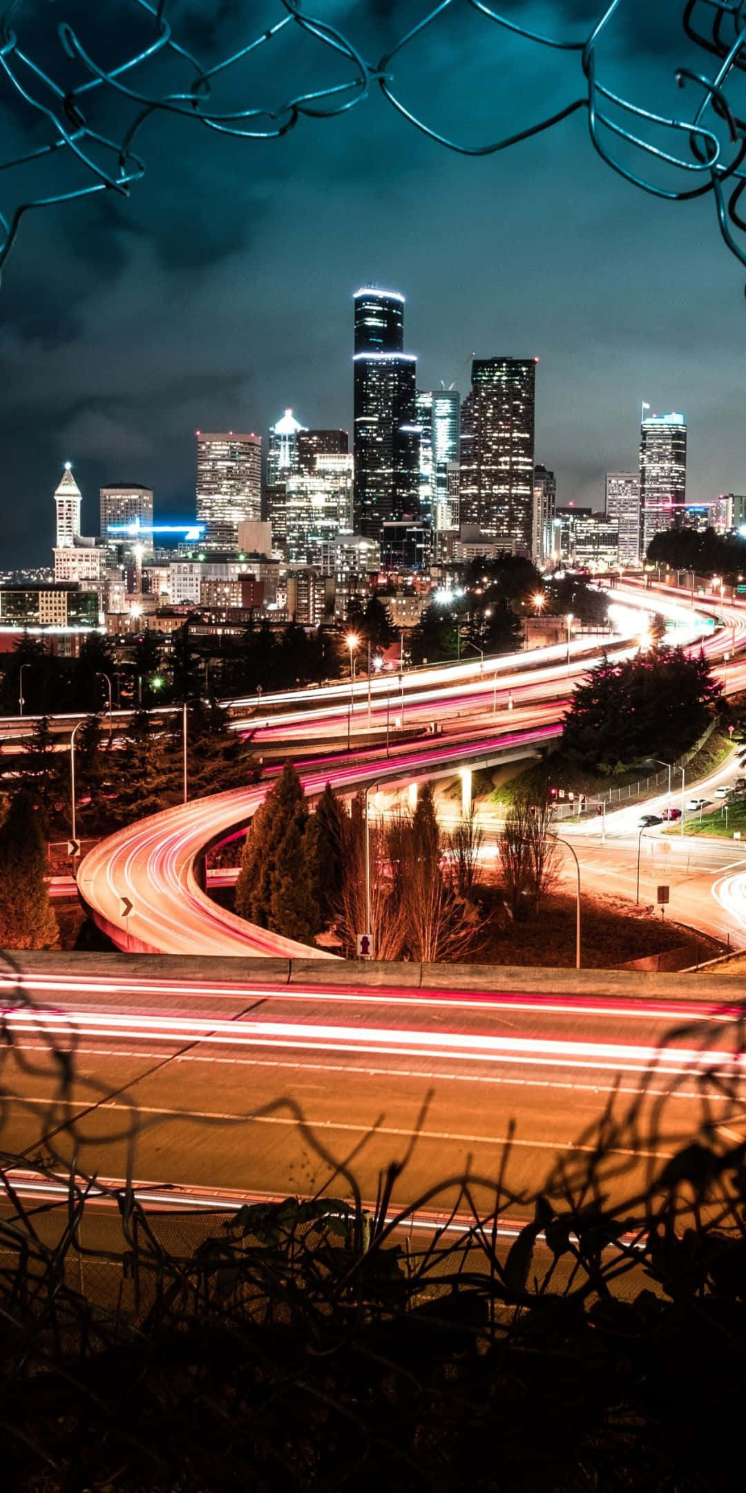 Pixel 3 Seattle 桌布 壁紙 — 夜晚城市景觀，透過鐵絲網圍欄看到發光的摩天大樓和車流的光跡。