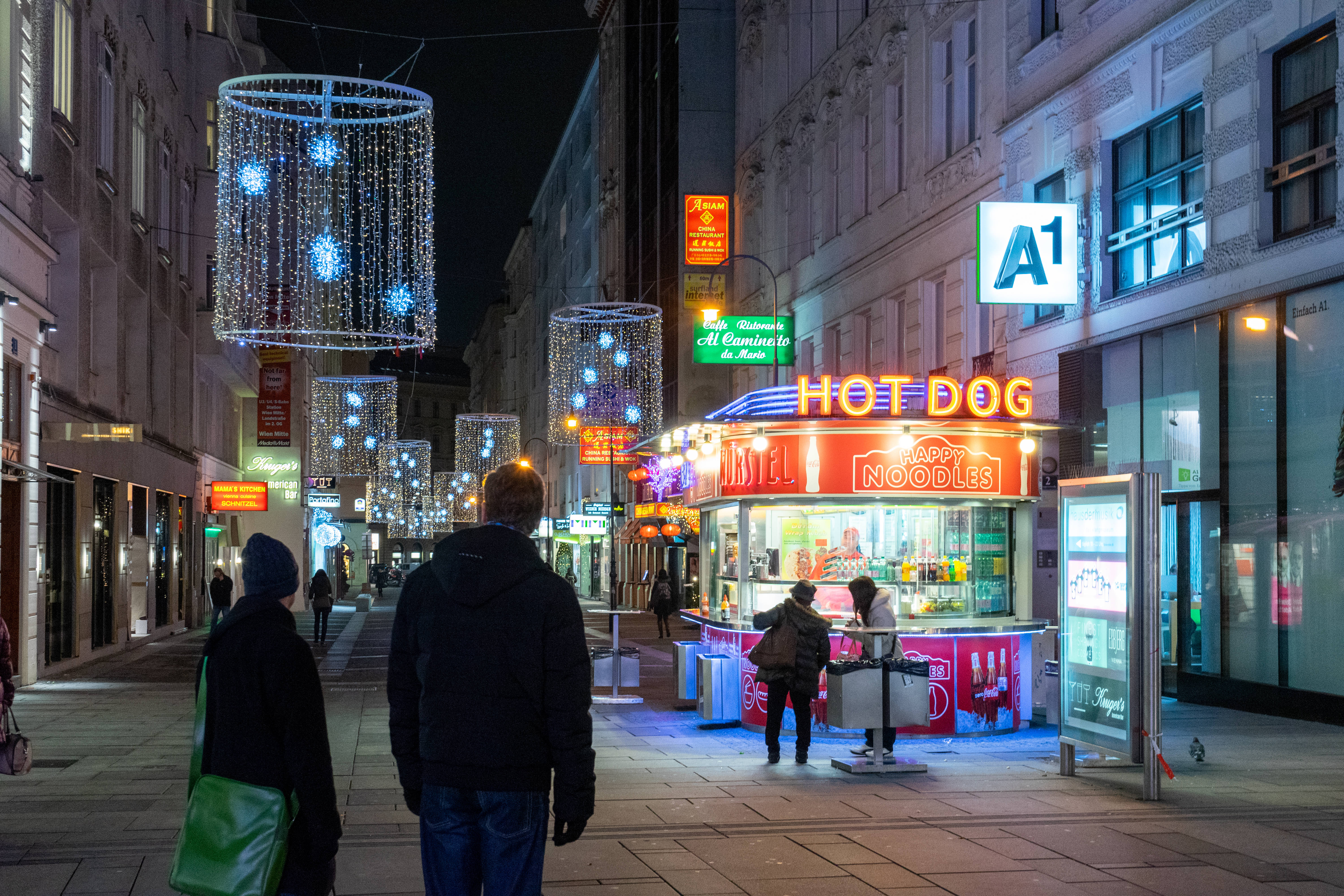 Austria 桌布 壁紙 — 夜晚城市街景，閃爍的霓虹燈招牌和懸掛的燈光裝置照亮熱狗攤附近的行人。