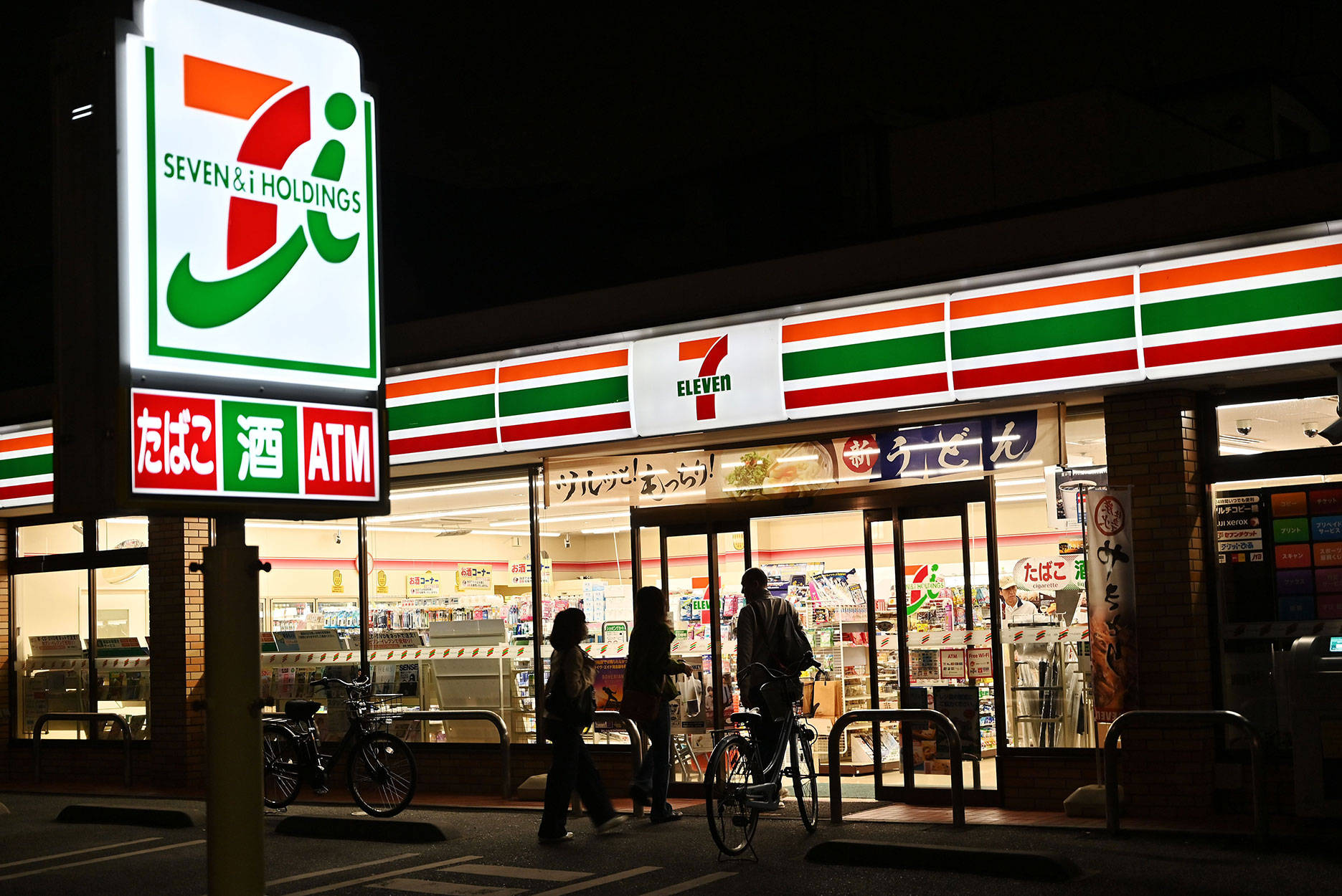 Eleven 桌布 壁紙 — 夜晚場景，一間燈火通明的7 Eleven便利商店，有行人和腳踏車經過。
