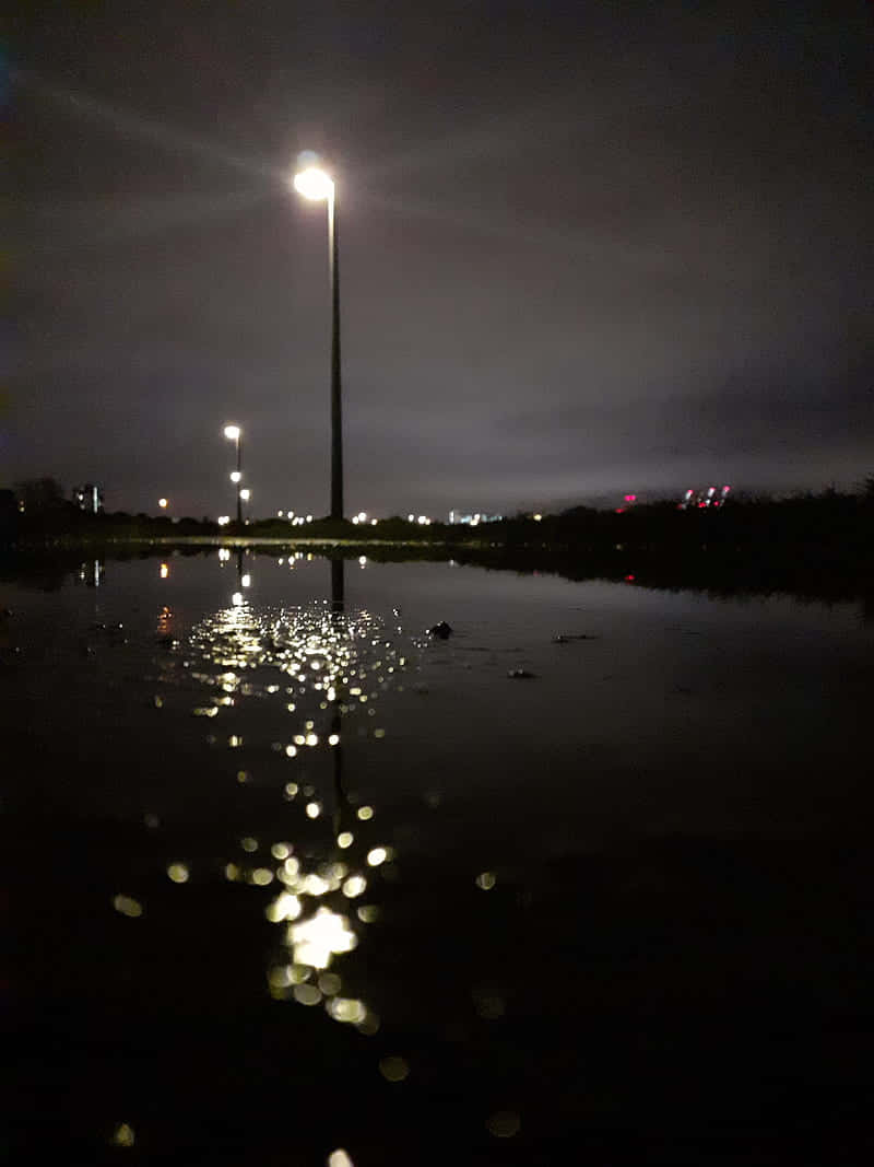 夜晚 桌布 壁紙 — 夜晚場景，高聳的路燈在平靜的水面上投下明亮的倒影，天空陰暗多雲。