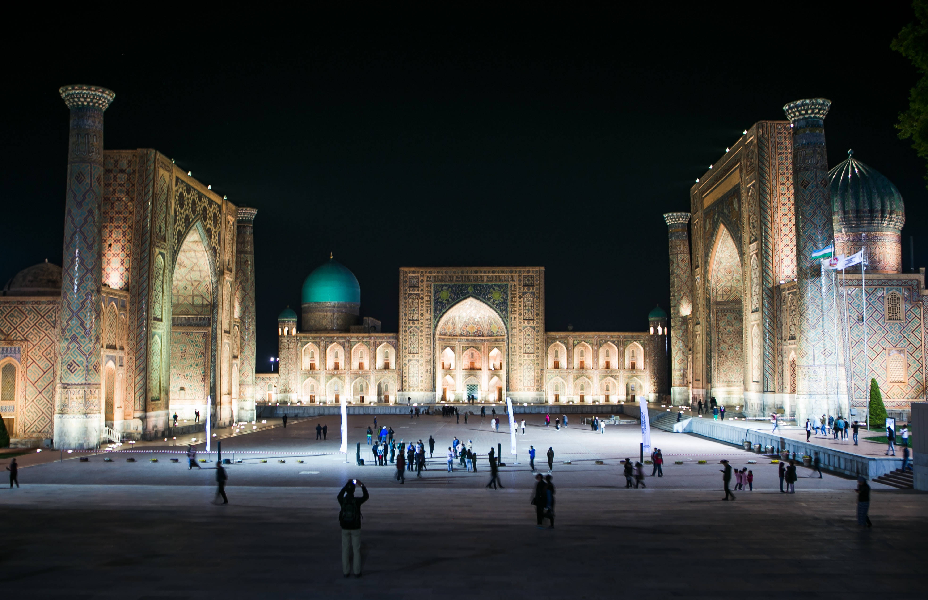 Uzbekistan 桌布 壁紙 — 夜晚宏偉的伊斯蘭建築，發光的圓頂和宣禮塔將溫暖的光線投射到充滿遊客的寬敞廣場上。