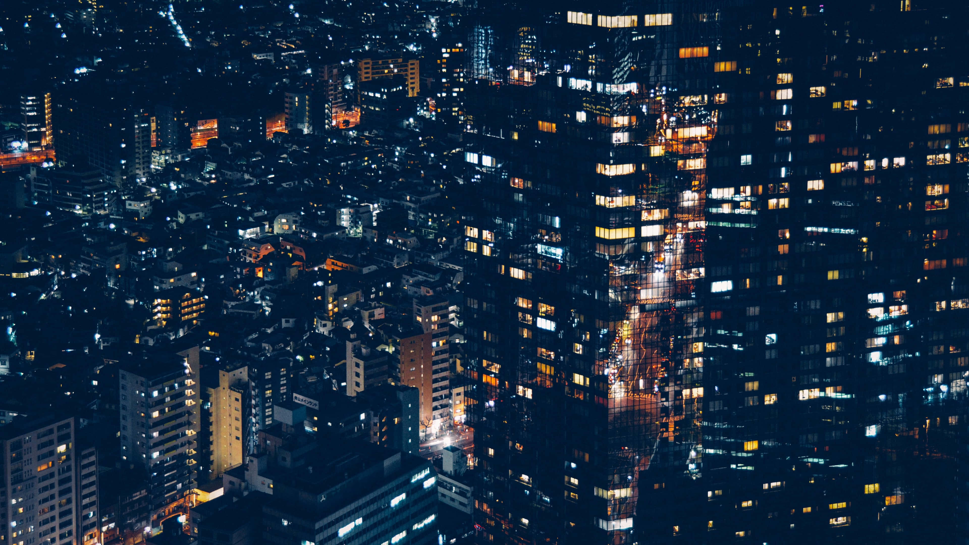 Harajuku 桌布 壁紙 — 夜晚密集城市的鳥瞰圖，發光的窗戶和照明街道創造出充滿活力的城市掛毯。