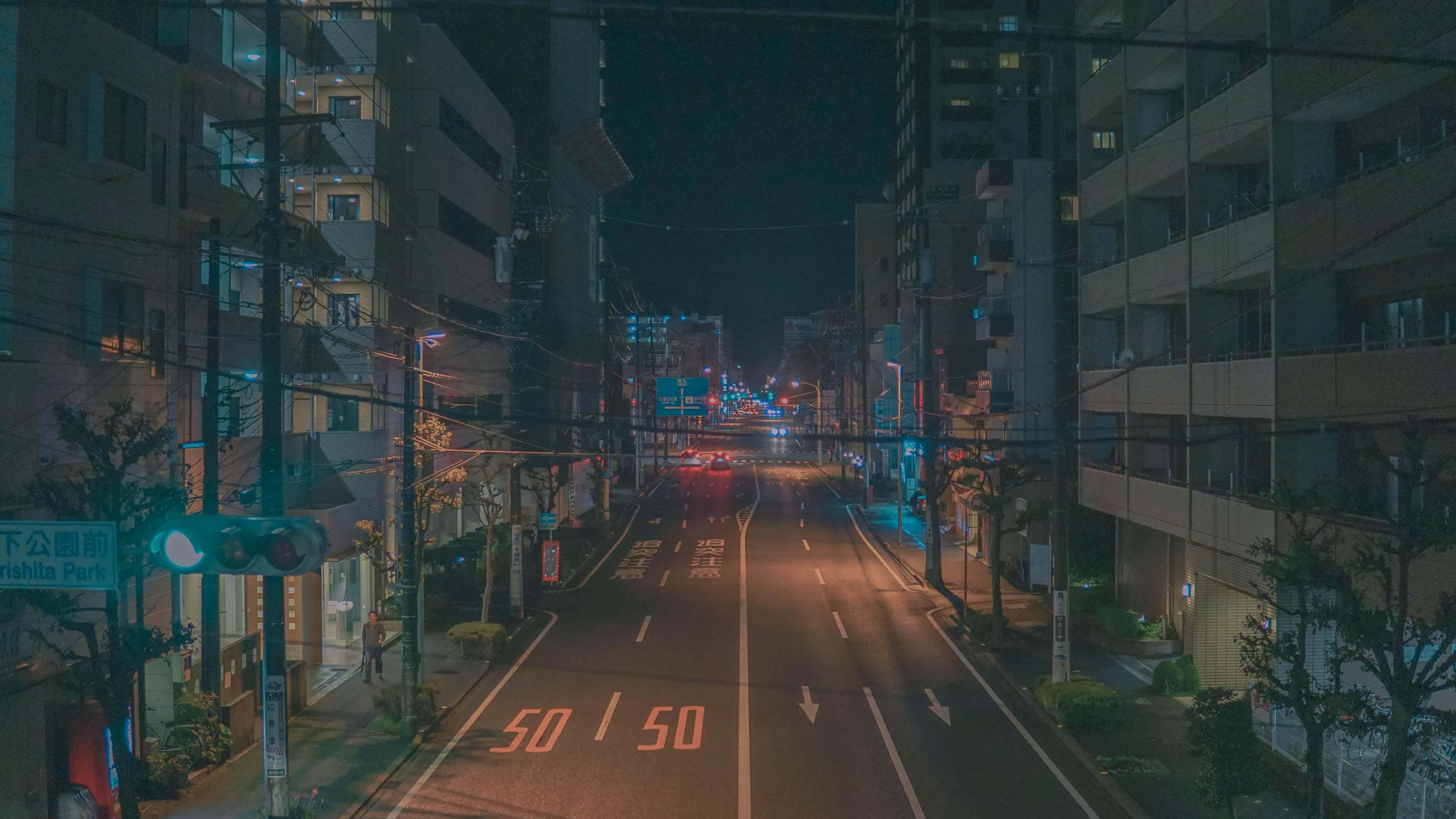 Cute Aesthetic Anime Desktop 桌布 壁紙 — 夜晚寧靜的城市街道，發光的街燈和遠處的車頭燈照亮了高樓之間的道路。