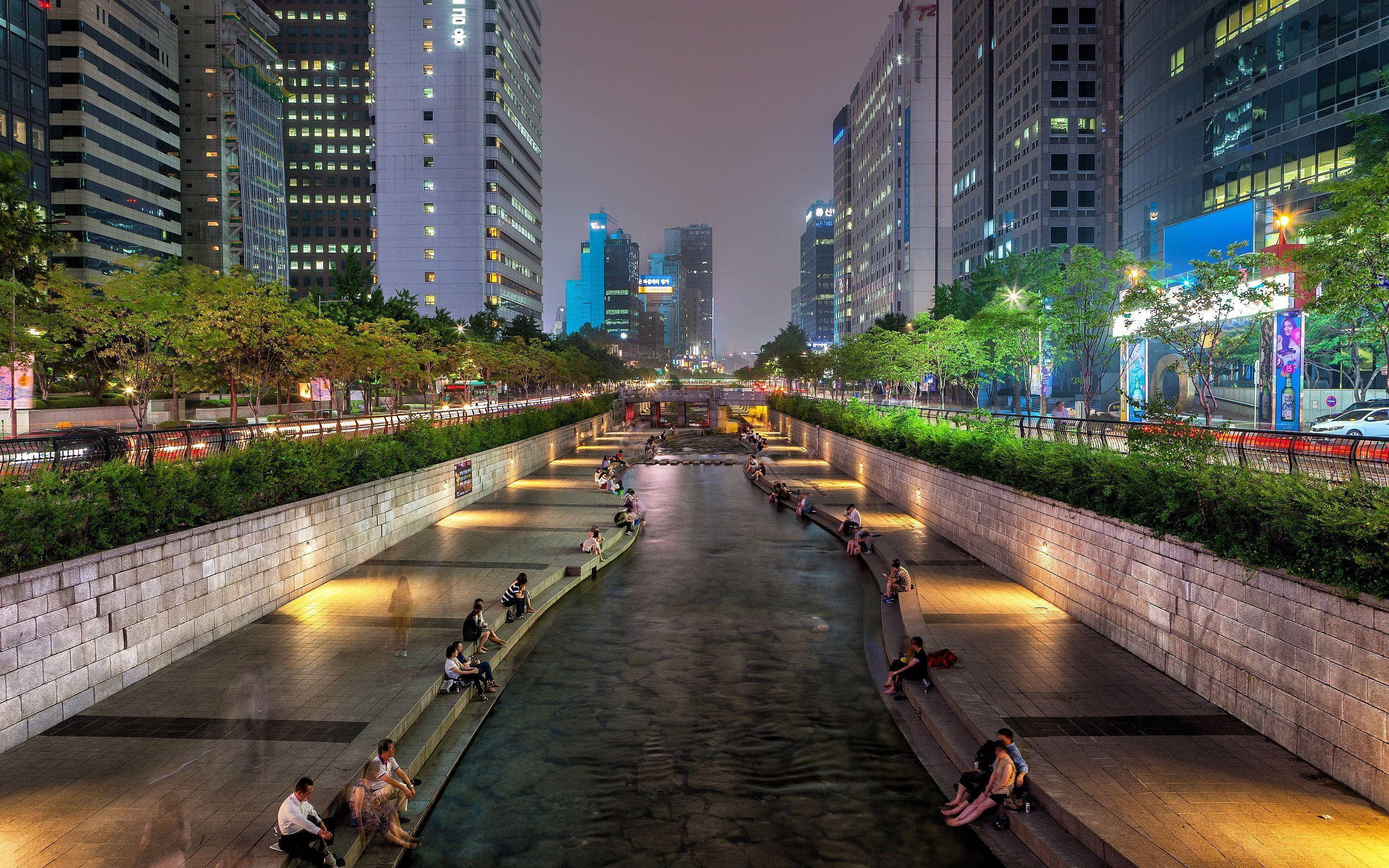 South Korea 桌布 壁紙 — 夜晚寧靜的城市運河，人們在溫暖燈光下的石凳上放鬆，兩旁是高聳的玻璃建築和綠植。