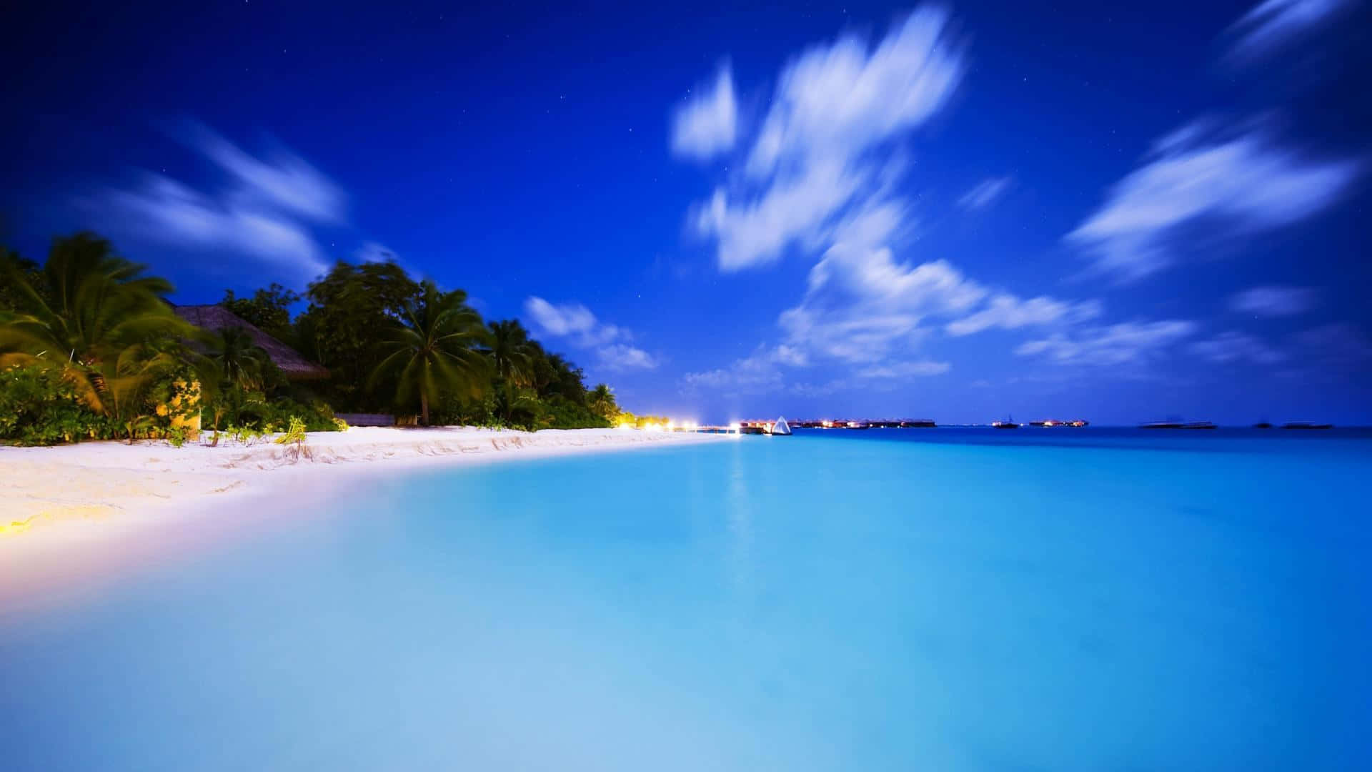 Ocean Beach 桌布 壁紙 — 夜晚寧靜的熱帶海灘，發光的藍色海水、棕櫚樹和滿天星斗的天空，伴隨著條紋狀的雲彩。
