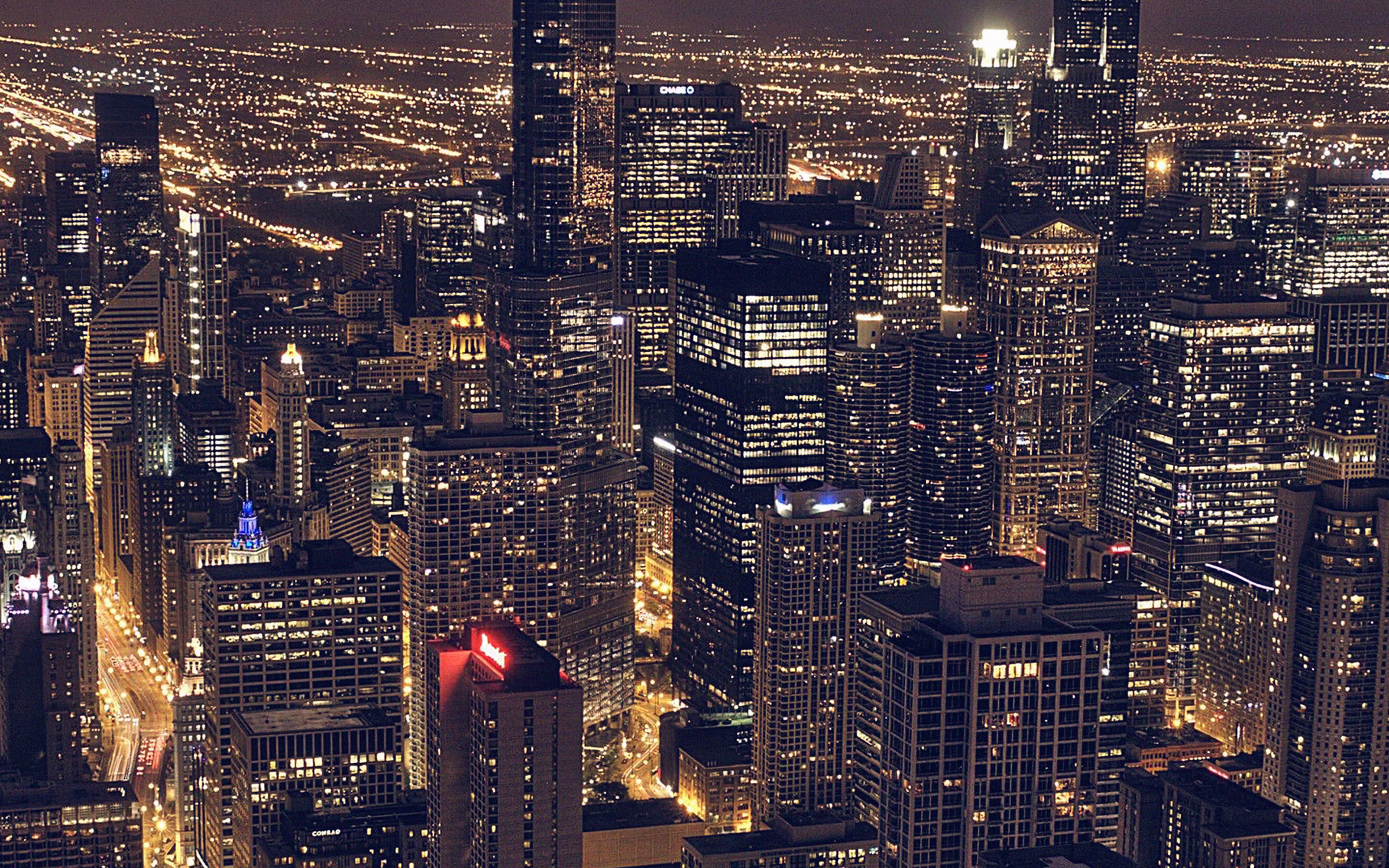 City Desktop 桌布 壁紙 — 夜晚廣闊城市景觀的鳥瞰圖，高聳的摩天大樓閃爍著溫暖的燈光，下方蜿蜒的街道燈火通明。