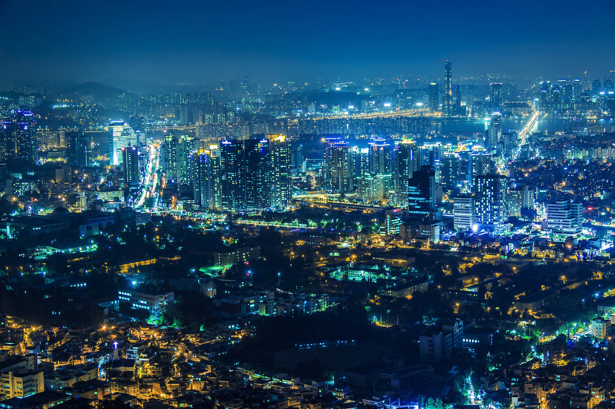 南韓 桌布 壁紙 — 夜晚廣闊的城市景觀，閃爍的摩天大樓和蜿蜒的街道被藍色和黃色的燈光照亮。