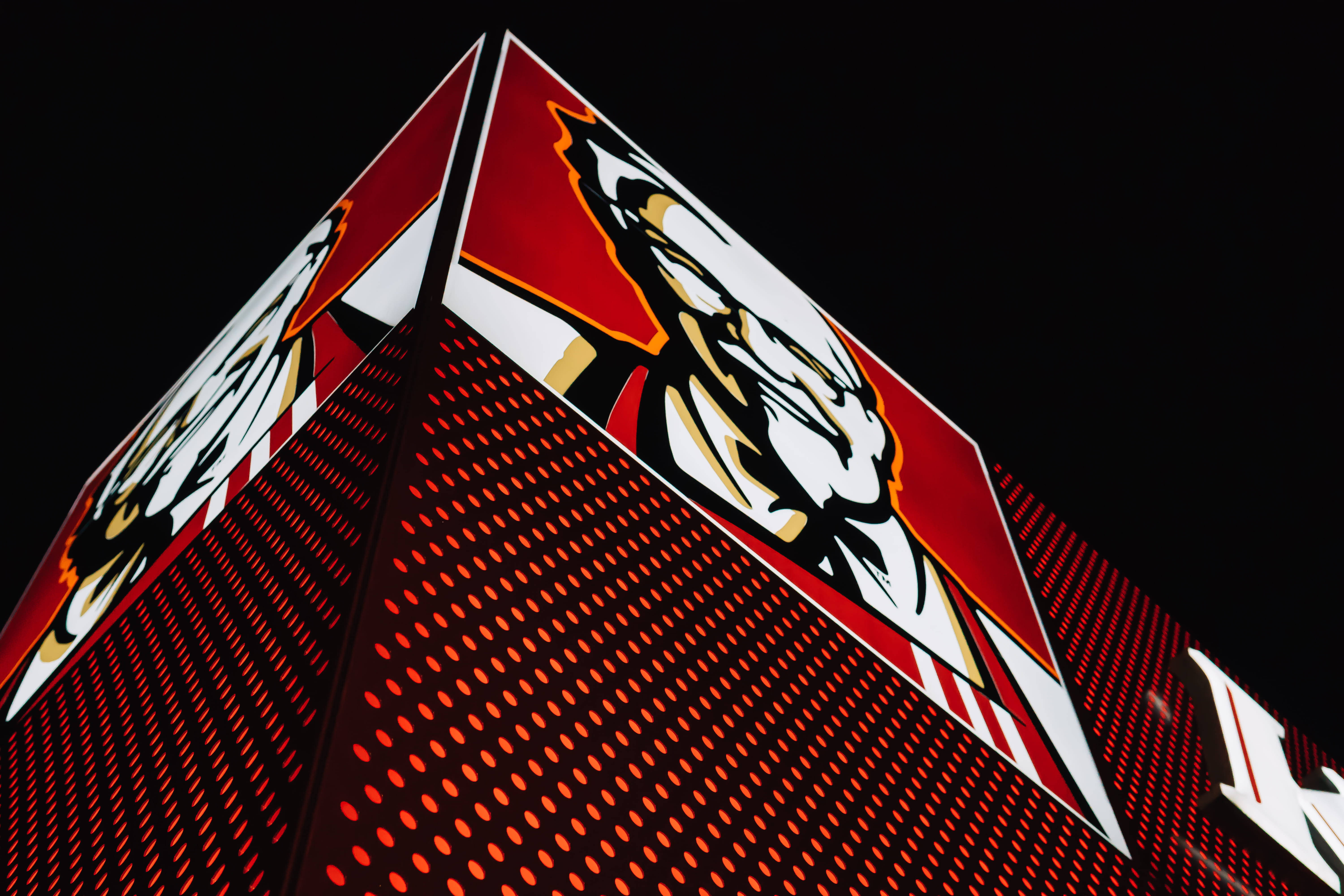 Kfc 桌布 壁紙 — 夜晚從低角度拍攝的金字塔形建築，其紅色穿孔外牆閃爍著內置燈光，並在黑色天空的襯托下飾有醒目的白色和金色虎頭標誌。