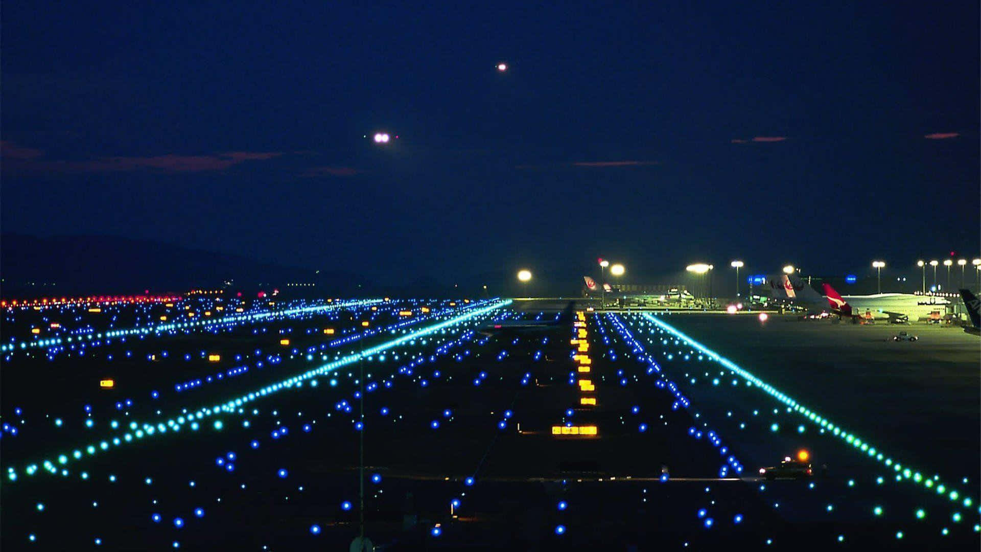 Runway 桌布 壁紙 — 夜晚從空中俯瞰機場跑道，在黑暗的天空下，藍色和黃色的燈光閃爍，遠處有飛機。