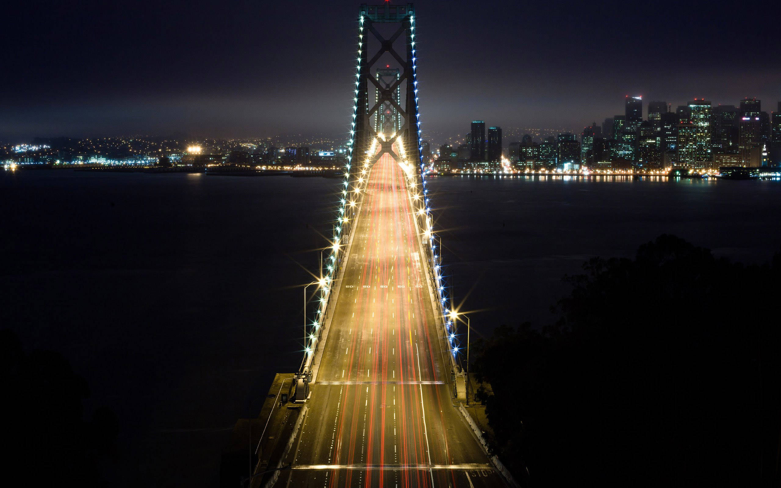 lights 桌布 壁紙 — 夜晚拍攝的長曝光照片，展示一座亮燈的吊橋，帶有流動的交通燈光和遠處發光的城市天際線。