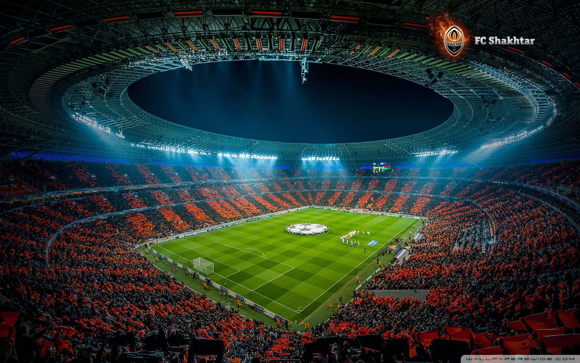 Soccer Stadium 桌布 壁紙 — 夜晚擠滿人的足球場鳥瞰圖，藍色聚光燈照亮了翠綠的球場，周圍是橙色球迷的海洋。