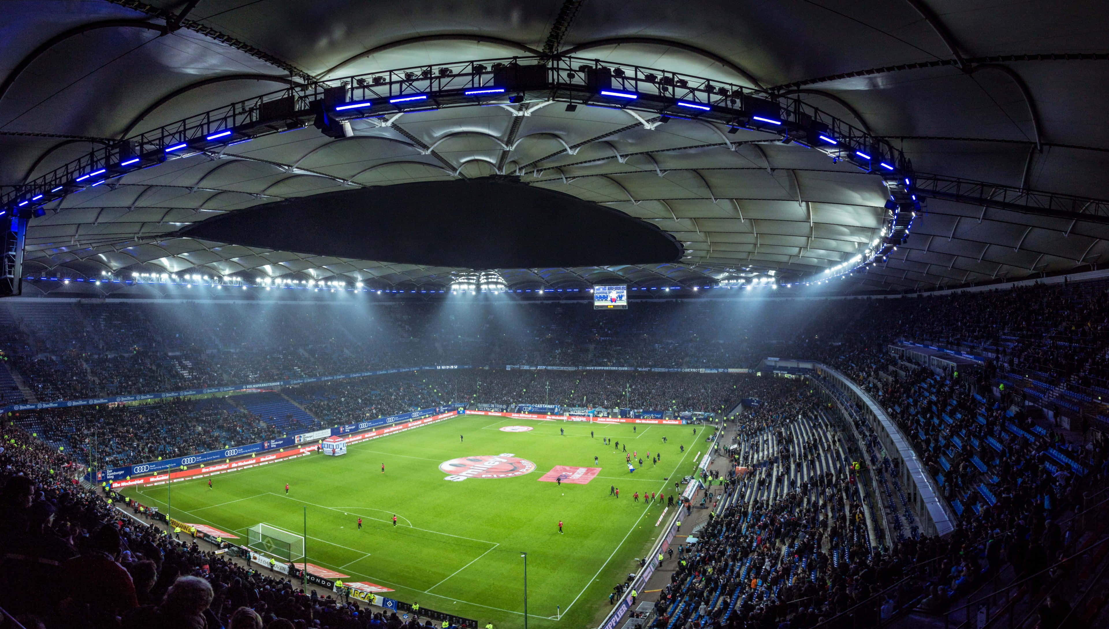 Soccer Stadium 桌布 壁紙 — 夜晚擠滿觀眾的足球場鳥瞰圖，明亮的泛光燈照亮弧形屋頂下的綠色球場。