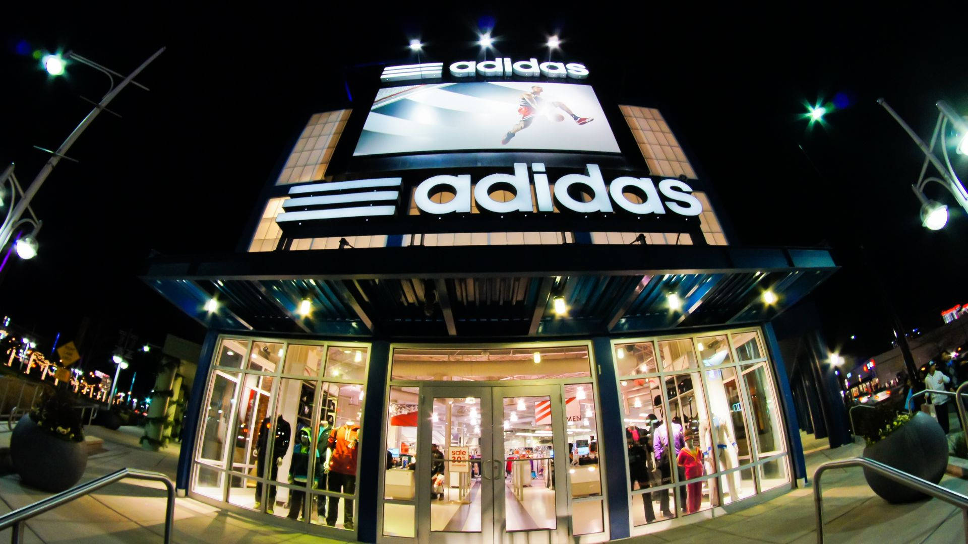 Clothes 桌布 壁紙 — 夜晚明亮照明的adidas商店，帶有發光標誌，透過玻璃窗可以看到裡面的人。